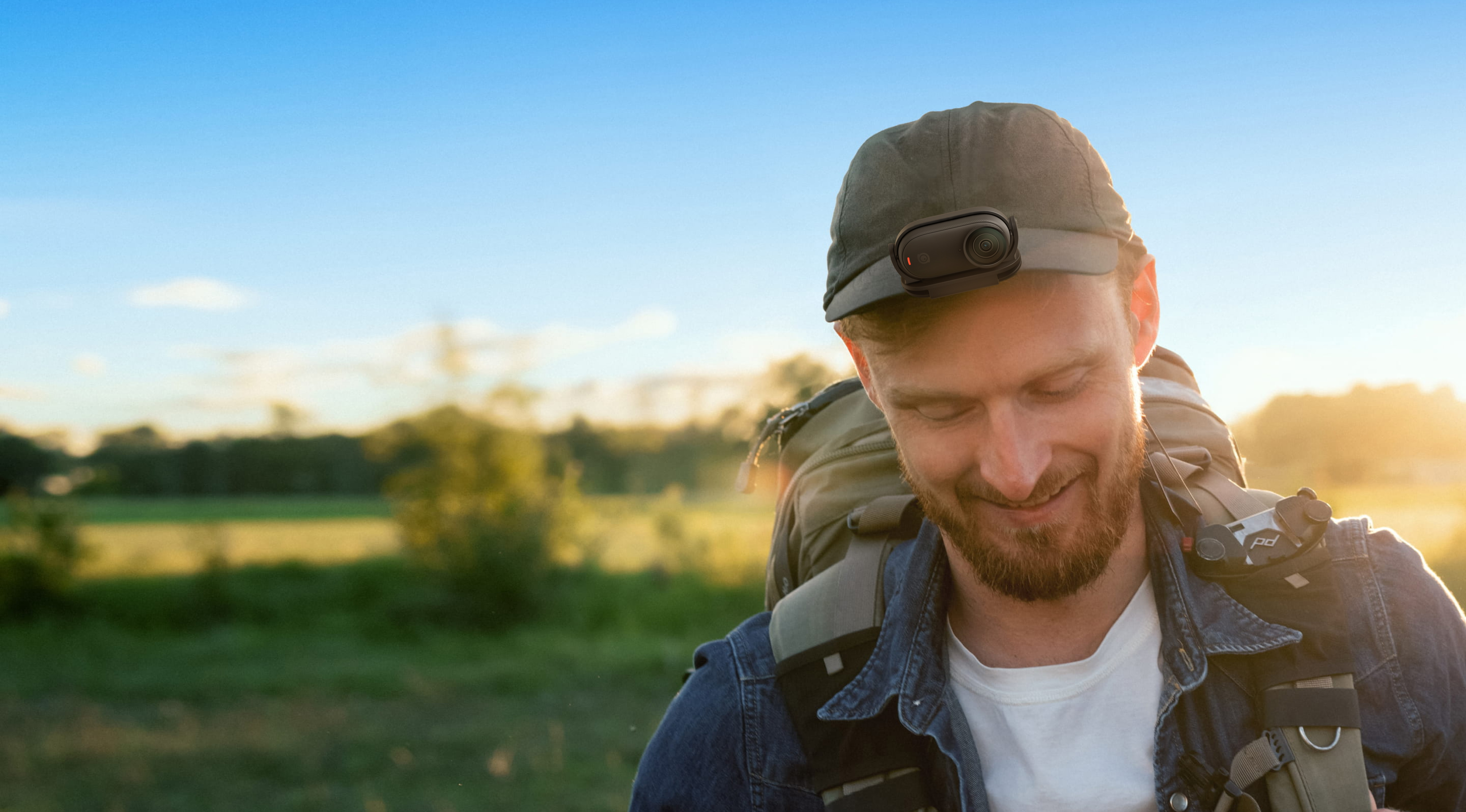 Mężczyzna z plecakiem i czapką z kamerą uśmiecha się. W tle pole.