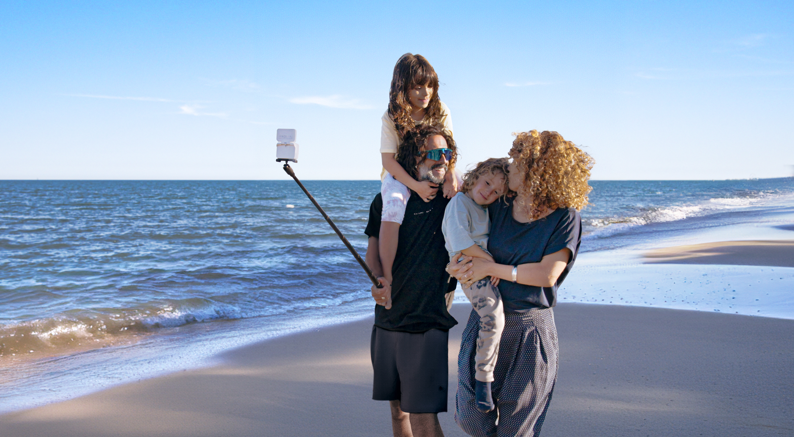 Rodzina na plaży pozuje do zdjęcia. Ojciec trzyma kij do selfie. Czyste, błękitne niebo.