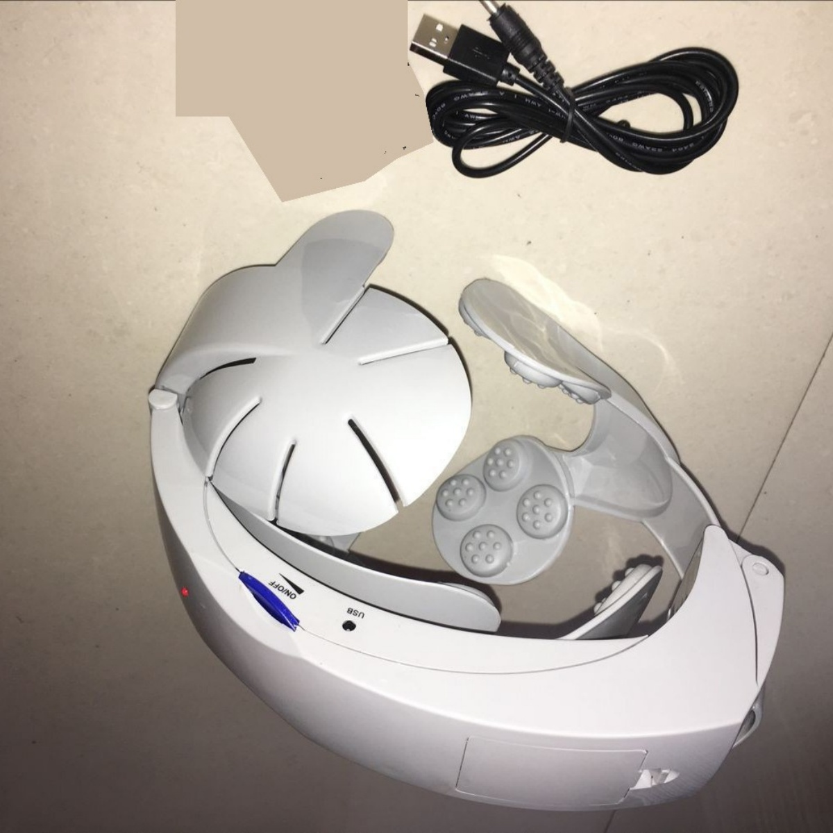 Weißes Kopfmassagegerät mit USB-Kabel. Hat eine gebogene Form und mehrere Massageknoten.