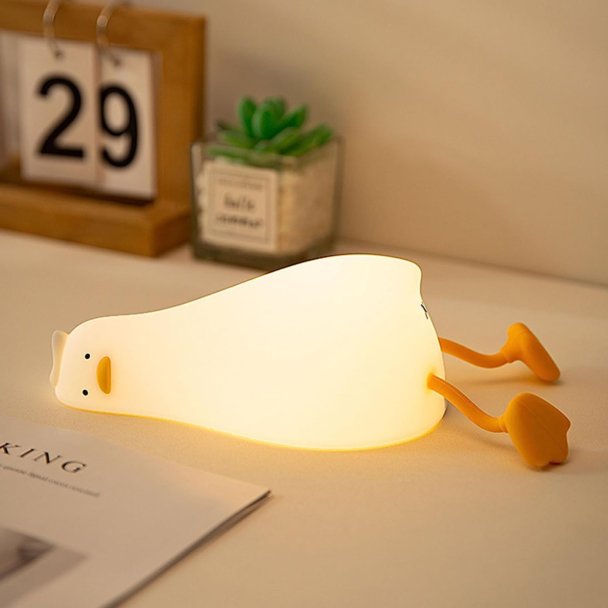 Entenförmige Nachtlampe mit weißem Körper, gelben Füßen und einem schwarzen Auge. Auf Tisch.