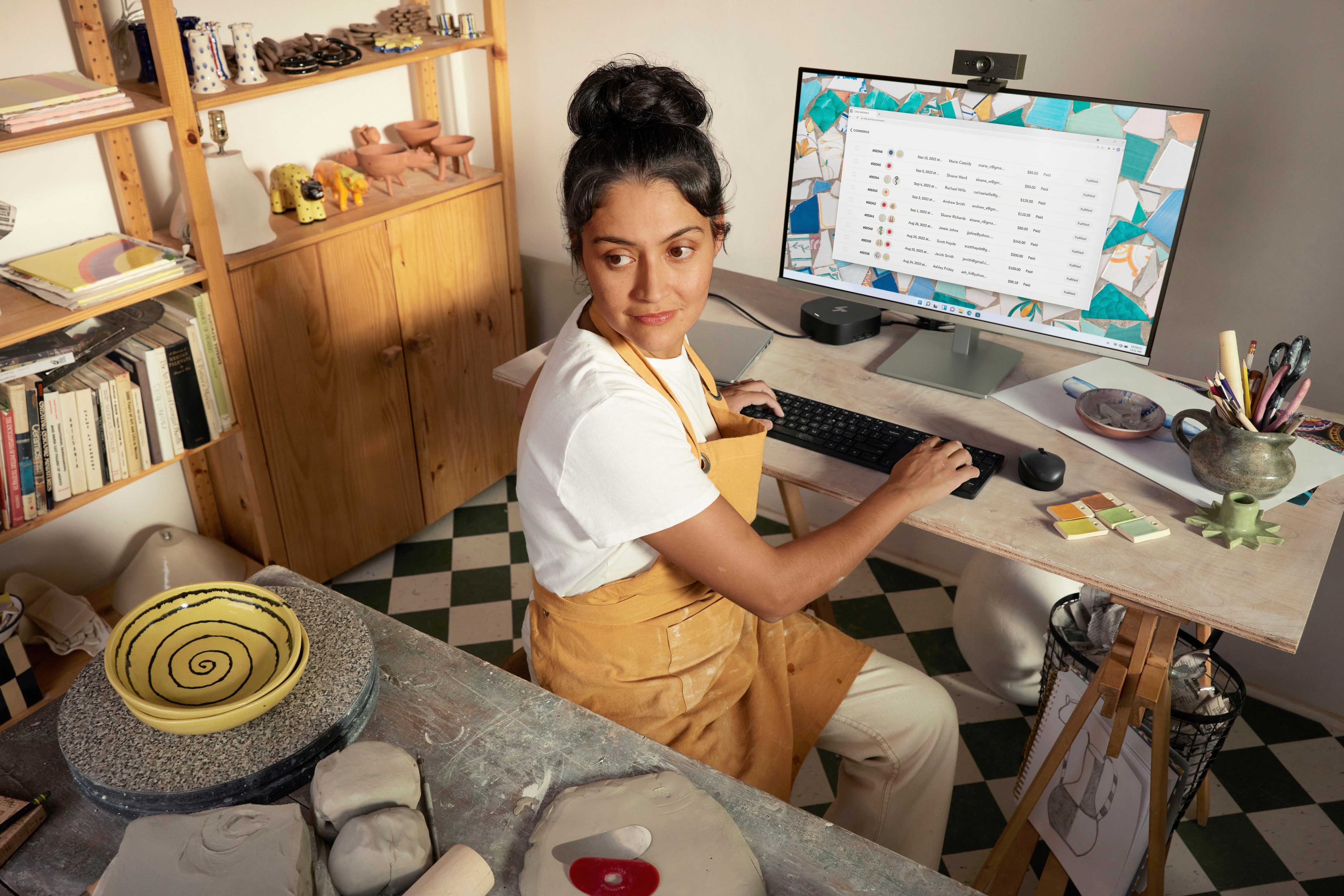 Kobieta przy biurku, pracująca na komputerze. Ceramika i materiały artystyczne w pobliżu. Szachownica na podłodze. Naturalne światło.