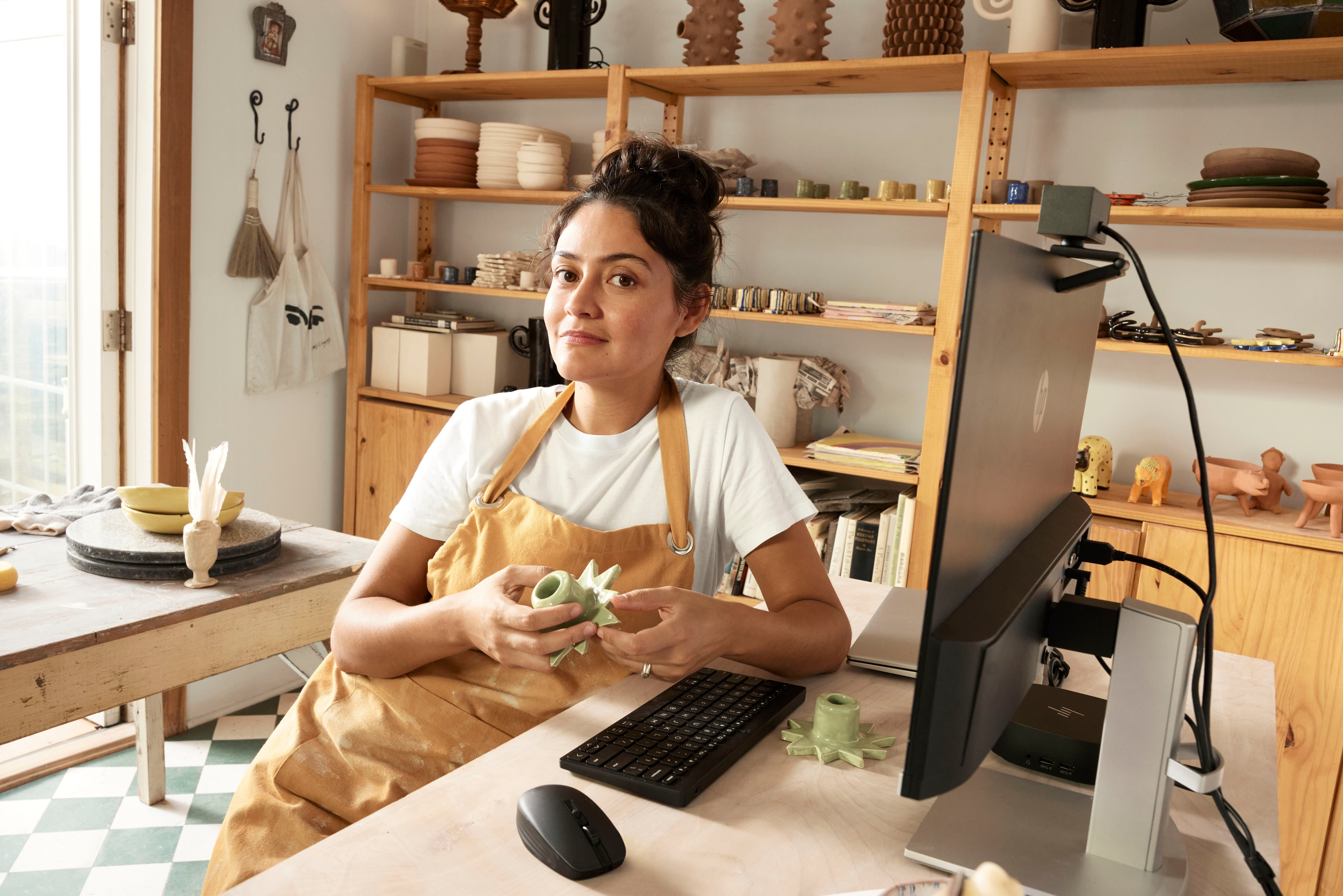Kobieta w fartuchu trzymająca zielony element ceramiczny, siedząca przy biurku z komputerem i akcesoriami do ceramiki.