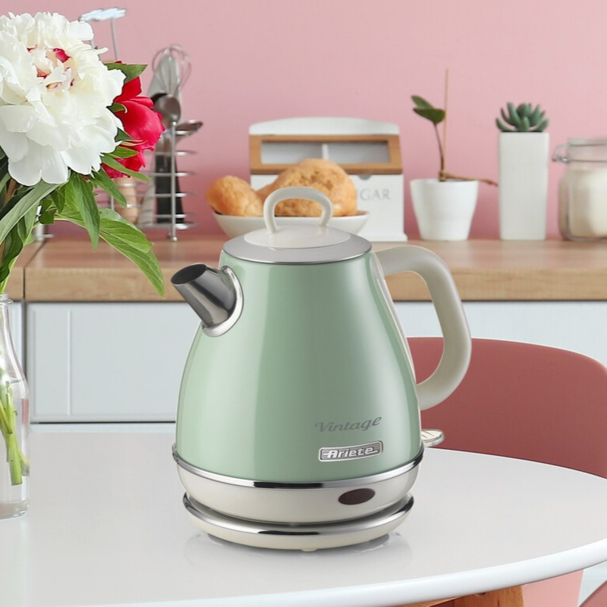 Miętowy czajnik Ariete Vintage z białym uchwytem i podstawą na białym stole. Kwiaty i różowa ściana.