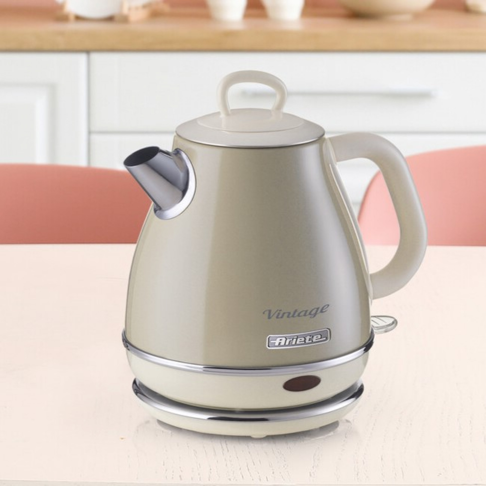 Beżowy czajnik elektryczny na białej podstawie. Stoi na drewnianej powierzchni, w pobliżu różowego krzesła.