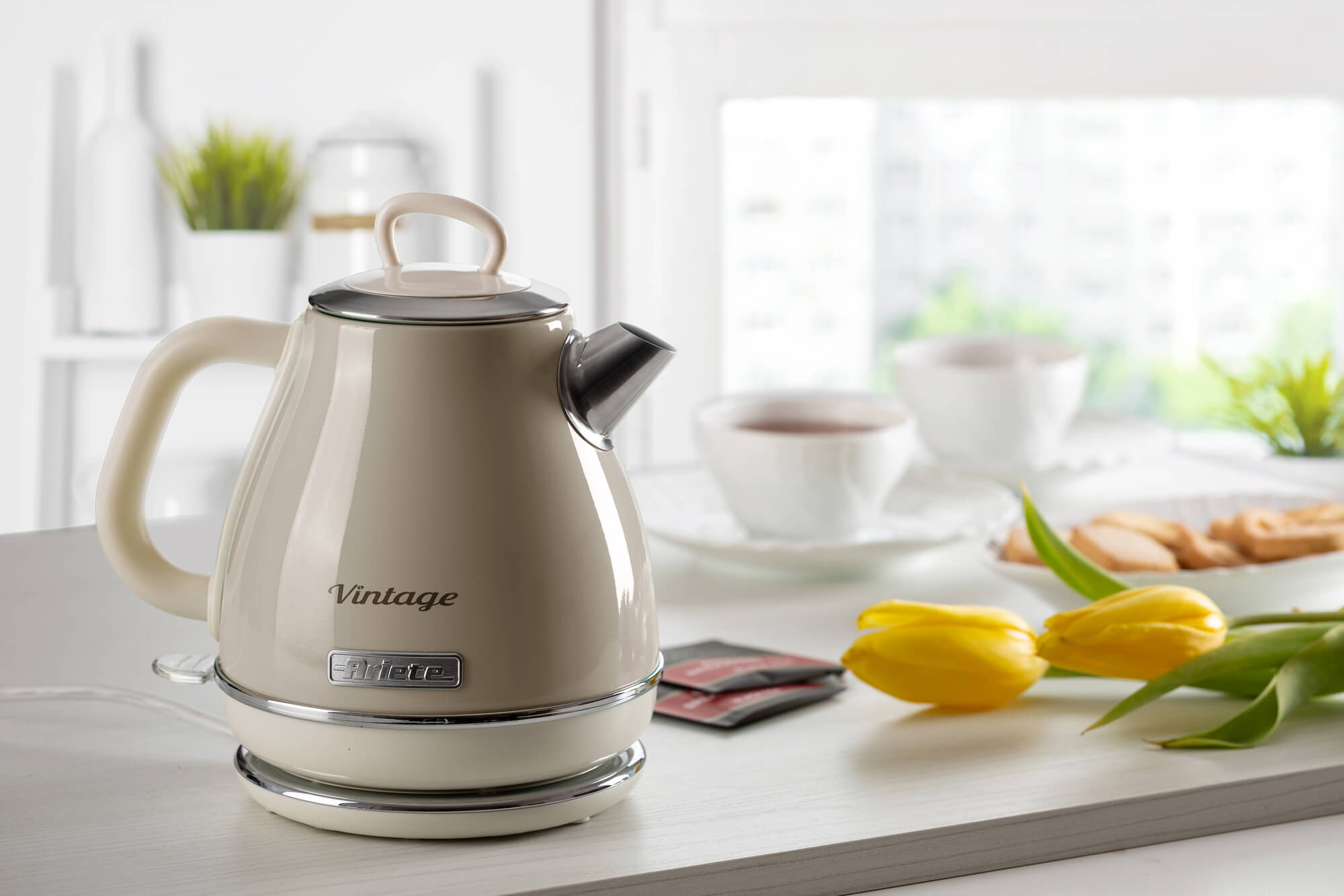 Beżowy czajnik elektryczny w stylu vintage, z wylewką, uchwytem i podstawą. W pobliżu dwa żółte tulipany.