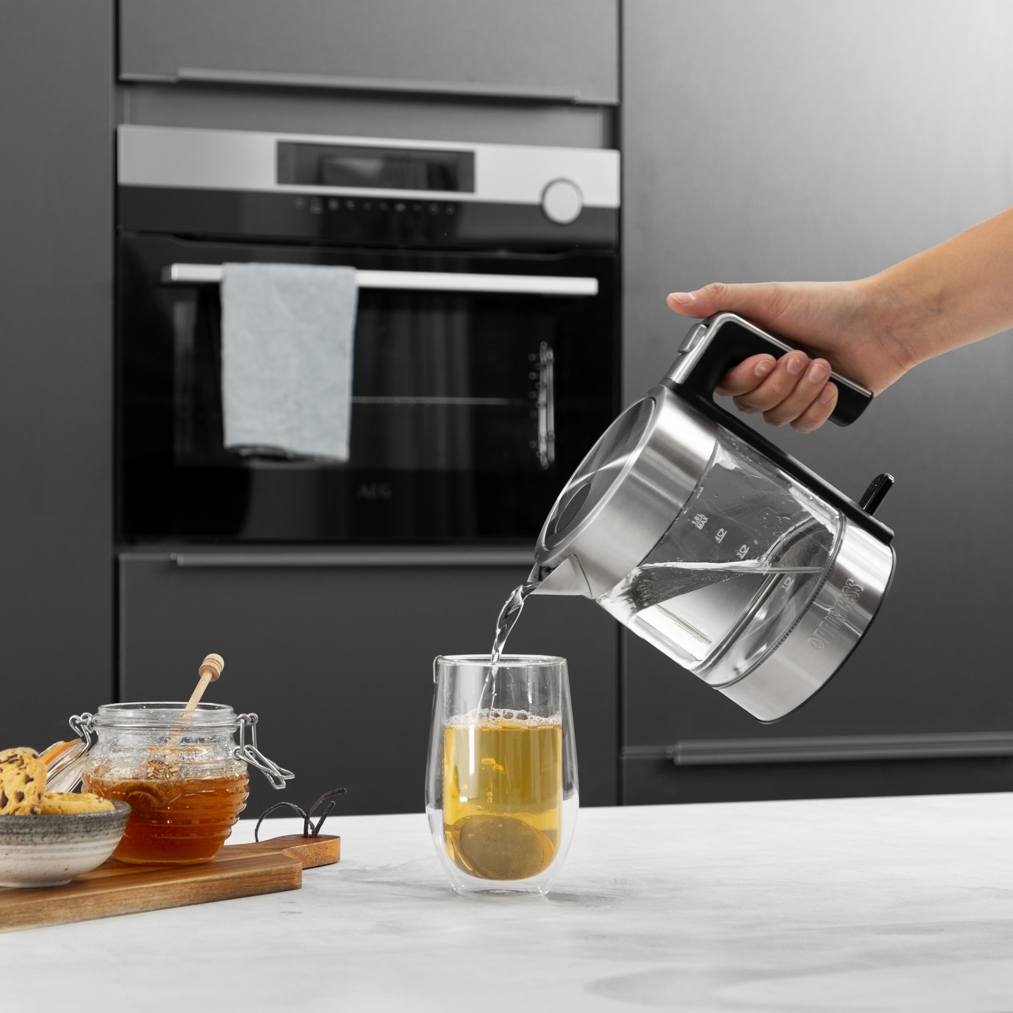 Osoba nalewa wodę z przezroczystego czajnika elektrycznego do szklanki herbaty z ciasteczkami w pobliżu.