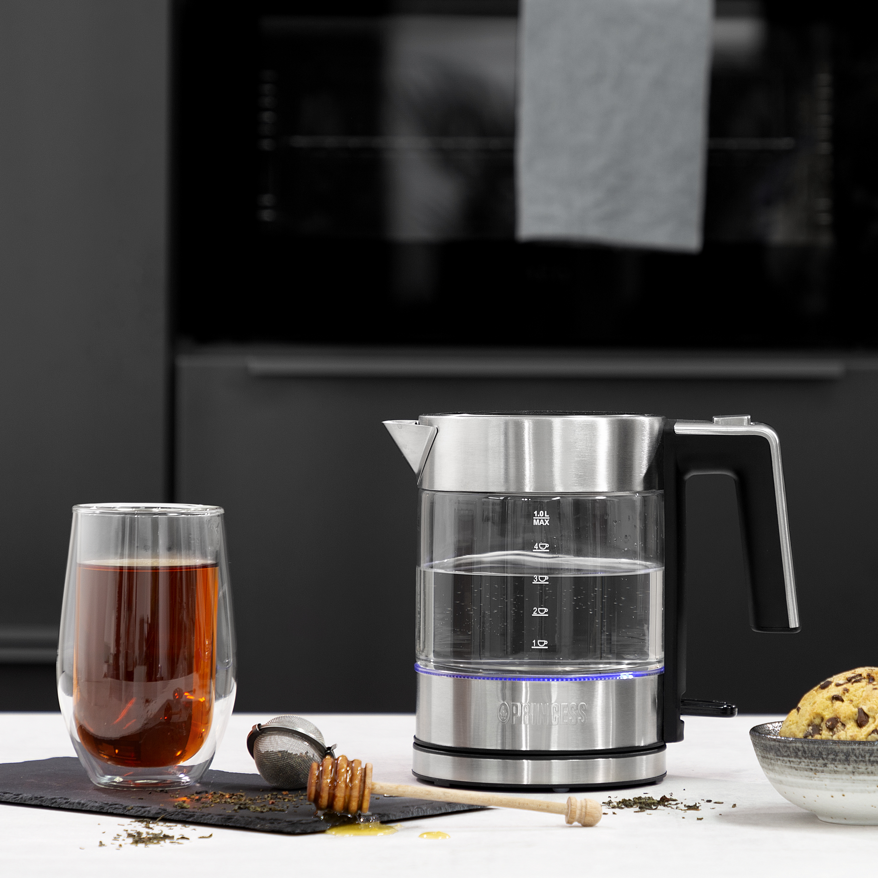 Szklany czajnik z czarnym uchwytem i podstawą. Jest szklanka herbaty i miska.