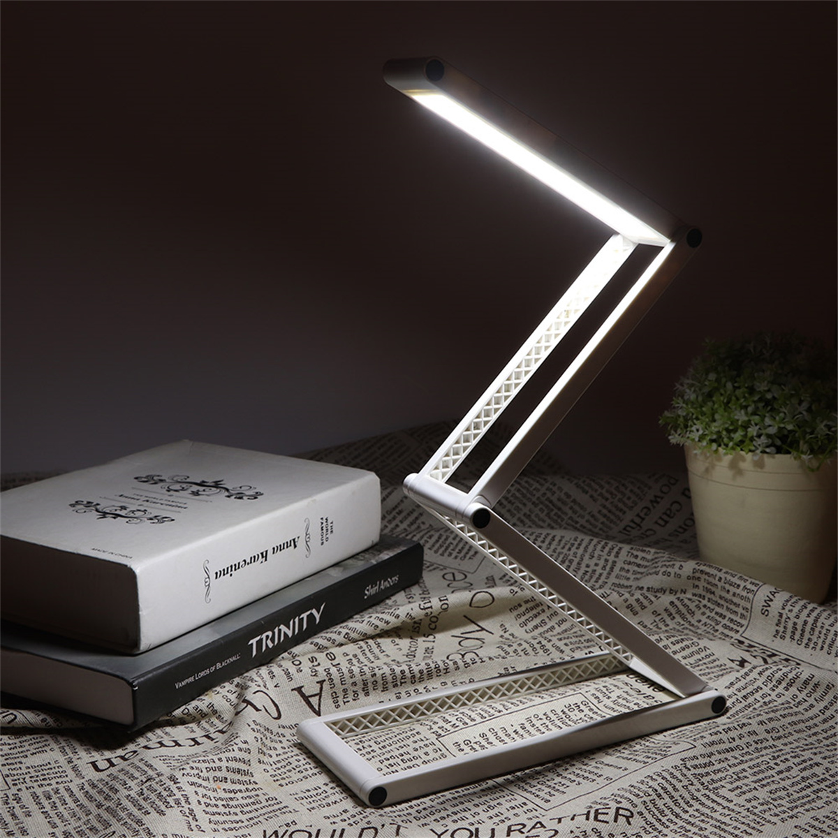 Faltbare weiße Schreibtischlampe mit hellem Licht auf einer gemusterten Oberfläche neben Büchern und einer Pflanze.