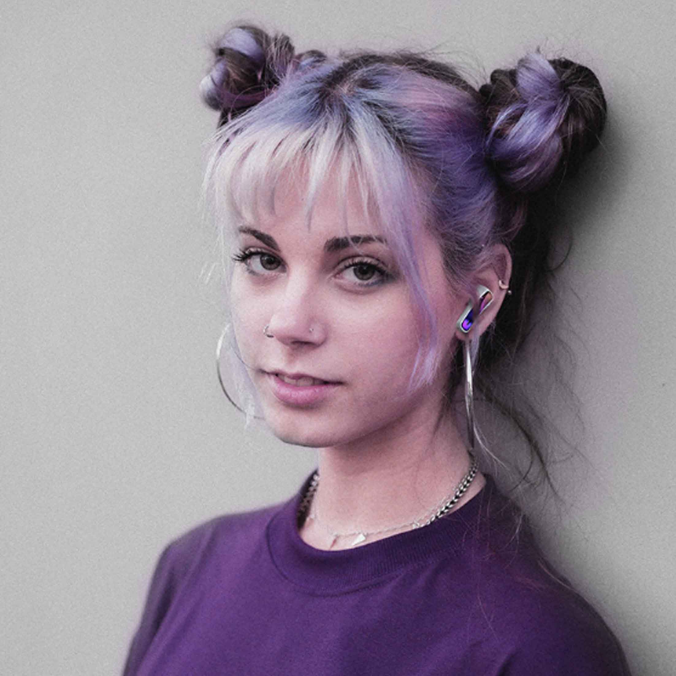 Junge Frau mit Space Buns, trägt Lila. Sie hat silberne Creolen und steht vor einer grauen Wand.