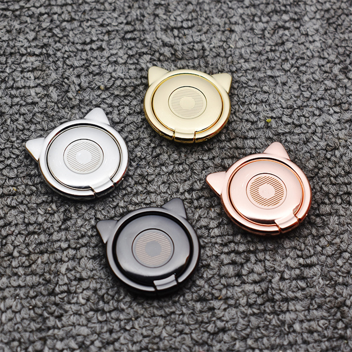 Vier katzenförmige Telefonhalter in Silber, Gold, Roségold und Schwarz auf grauer Oberfläche.