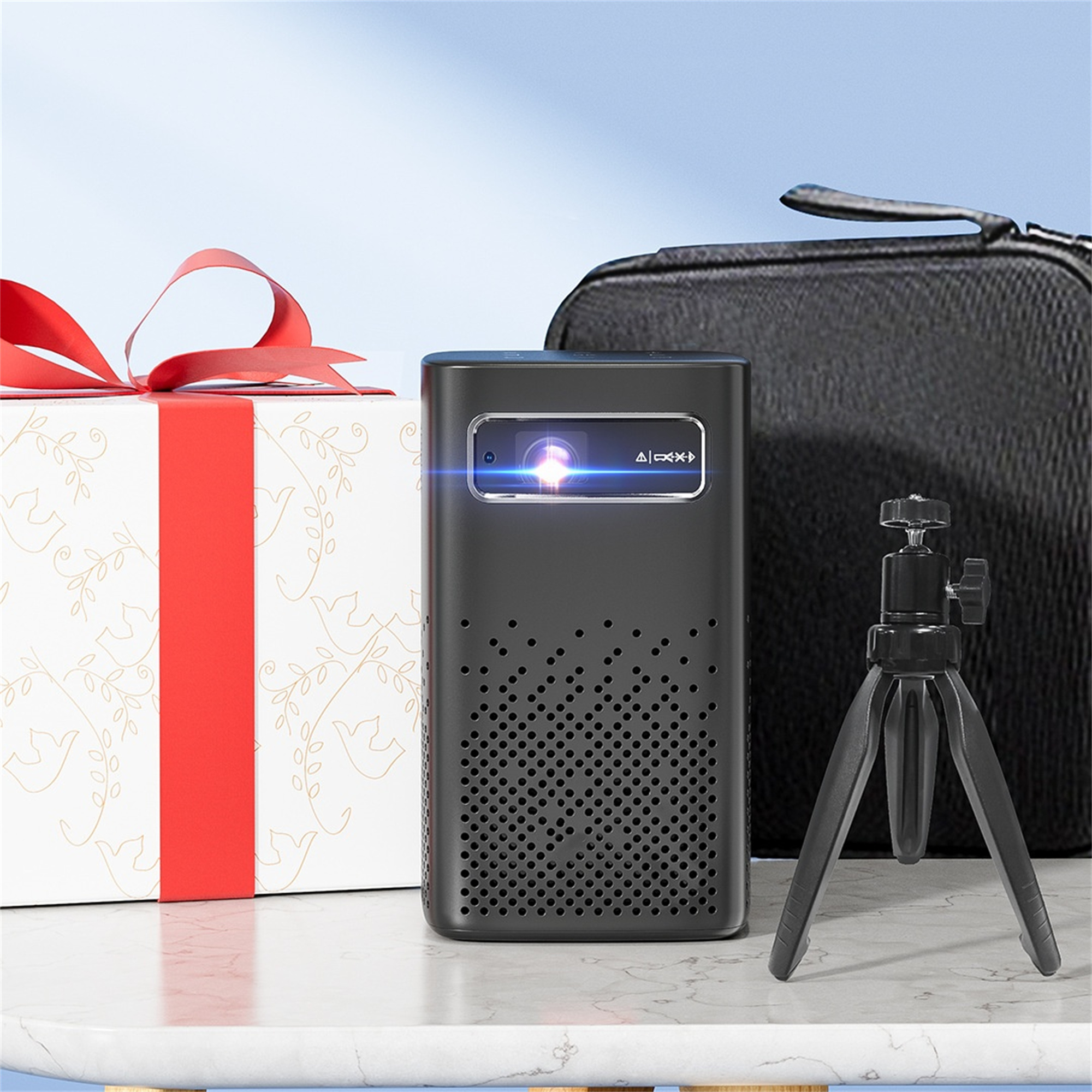 Schwarzer Projektor mit Geschenkbox, Stativ und Tragetasche. Weißer Tisch, blauer Hintergrund.