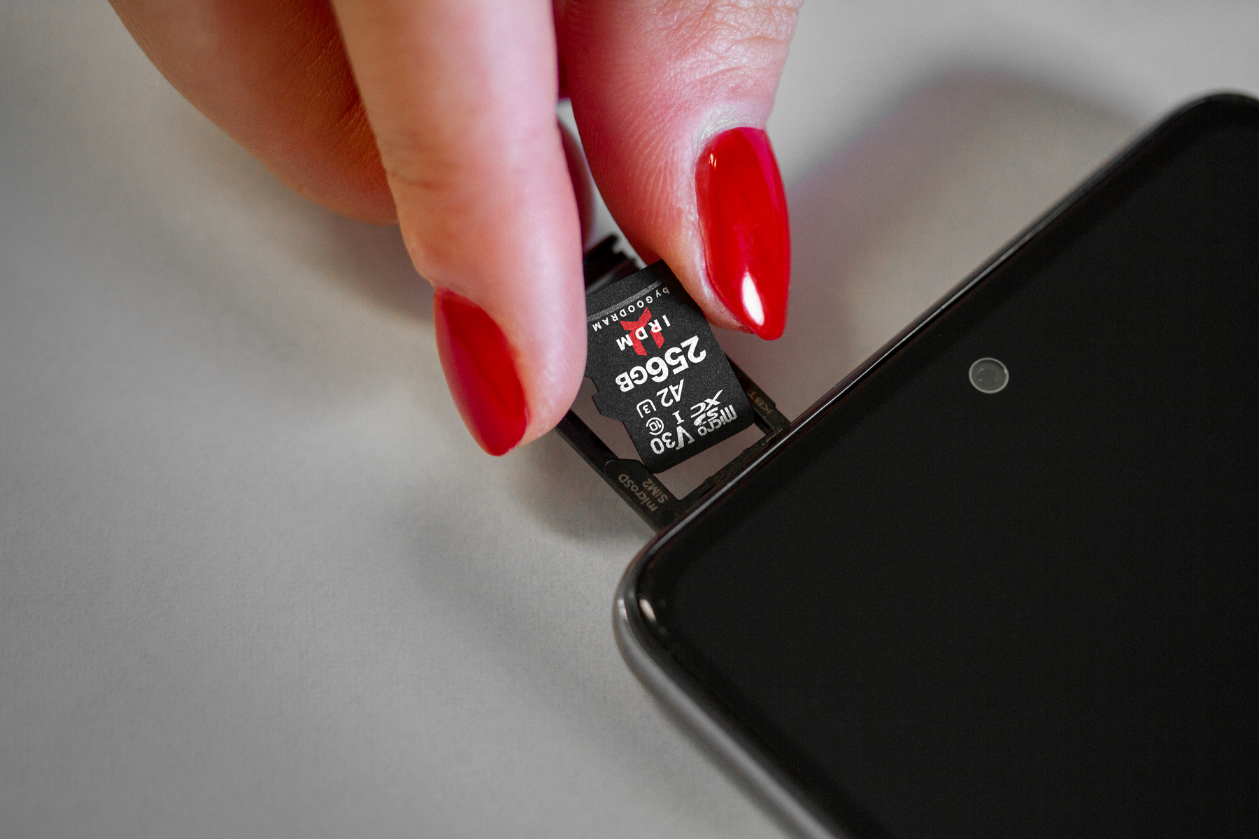 Dłoń z czerwonymi paznokciami wkładająca kartę microSD 256GB do telefonu. Białe tło.