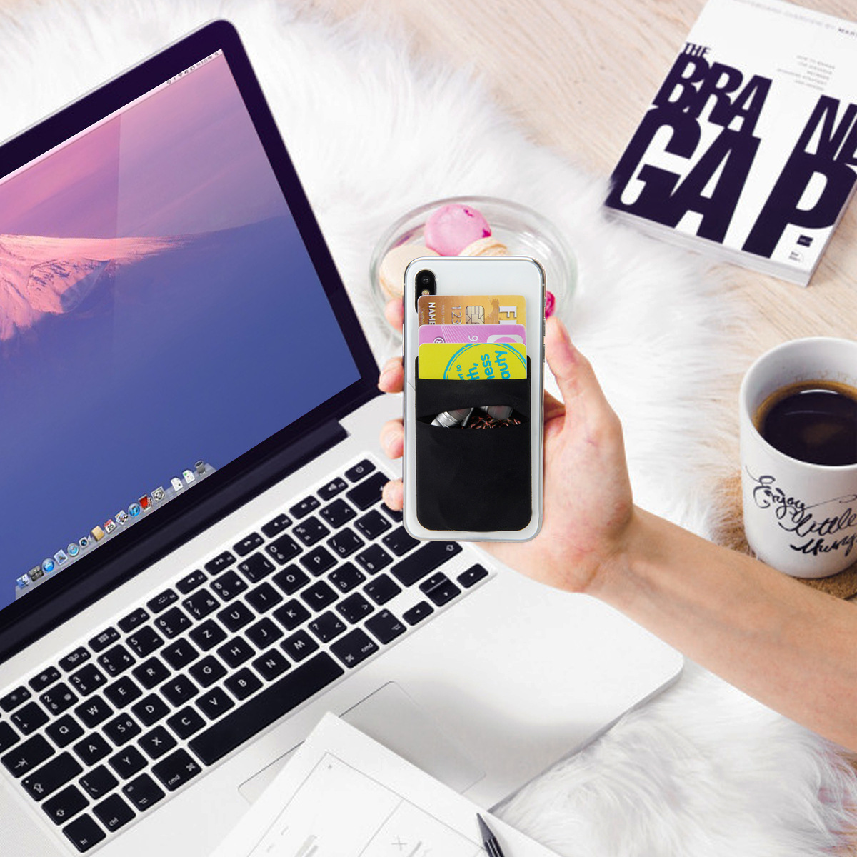 Dłoń trzyma etui na telefon z kartami i telefonem w środku. Widoczny laptop, książka i kubek.