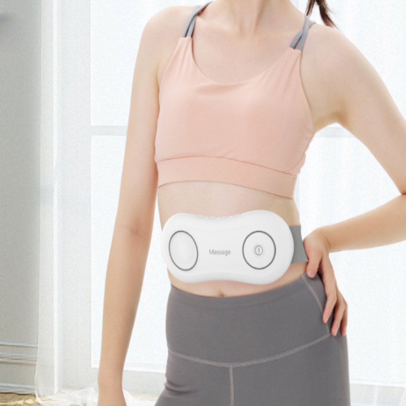 Frau mit einem weißen Massagegerät am Bauch. Sie trägt einen Sport-BH und graue Hose.