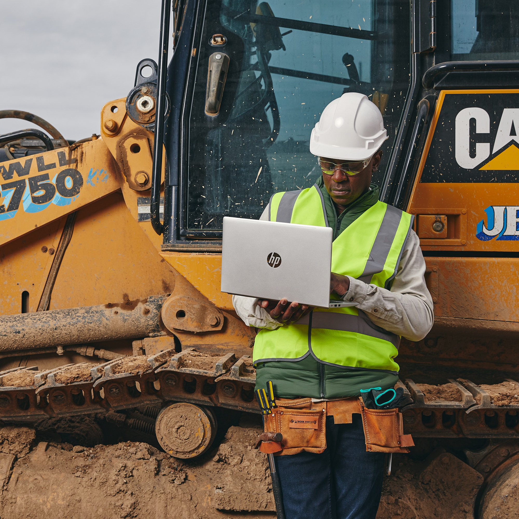 Operaio edile con laptop in piedi vicino a un bulldozer, con casco e giubbotto.