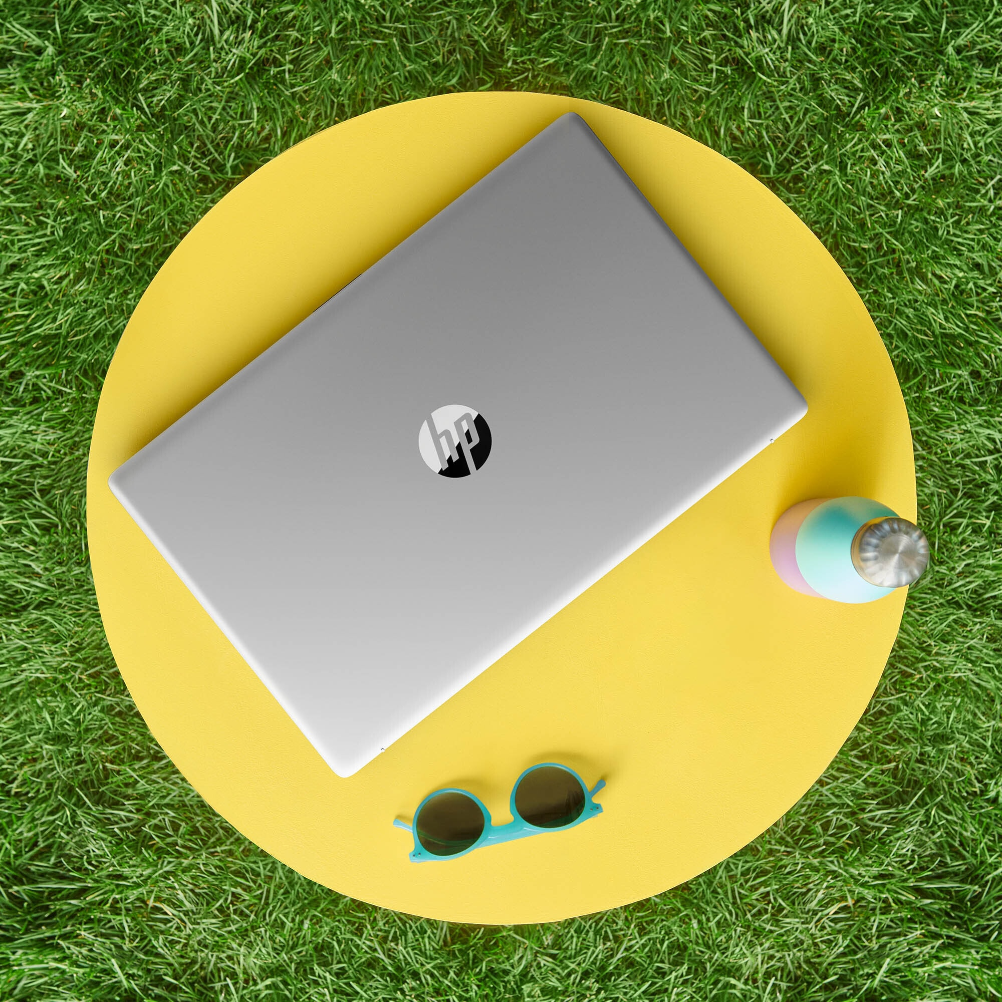 Laptop auf gelbem Tisch, Sonnenbrille und Wasserflasche auf Gras, Draufsicht.