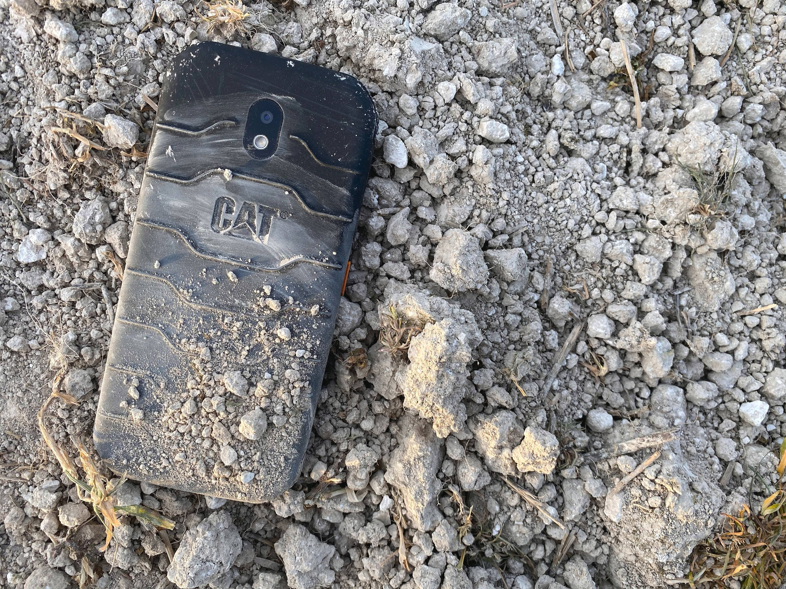 Schwarzes robustes CAT-Telefon liegt in Schmutz und kleinen Steinen. Das Telefon ist schmutzig.