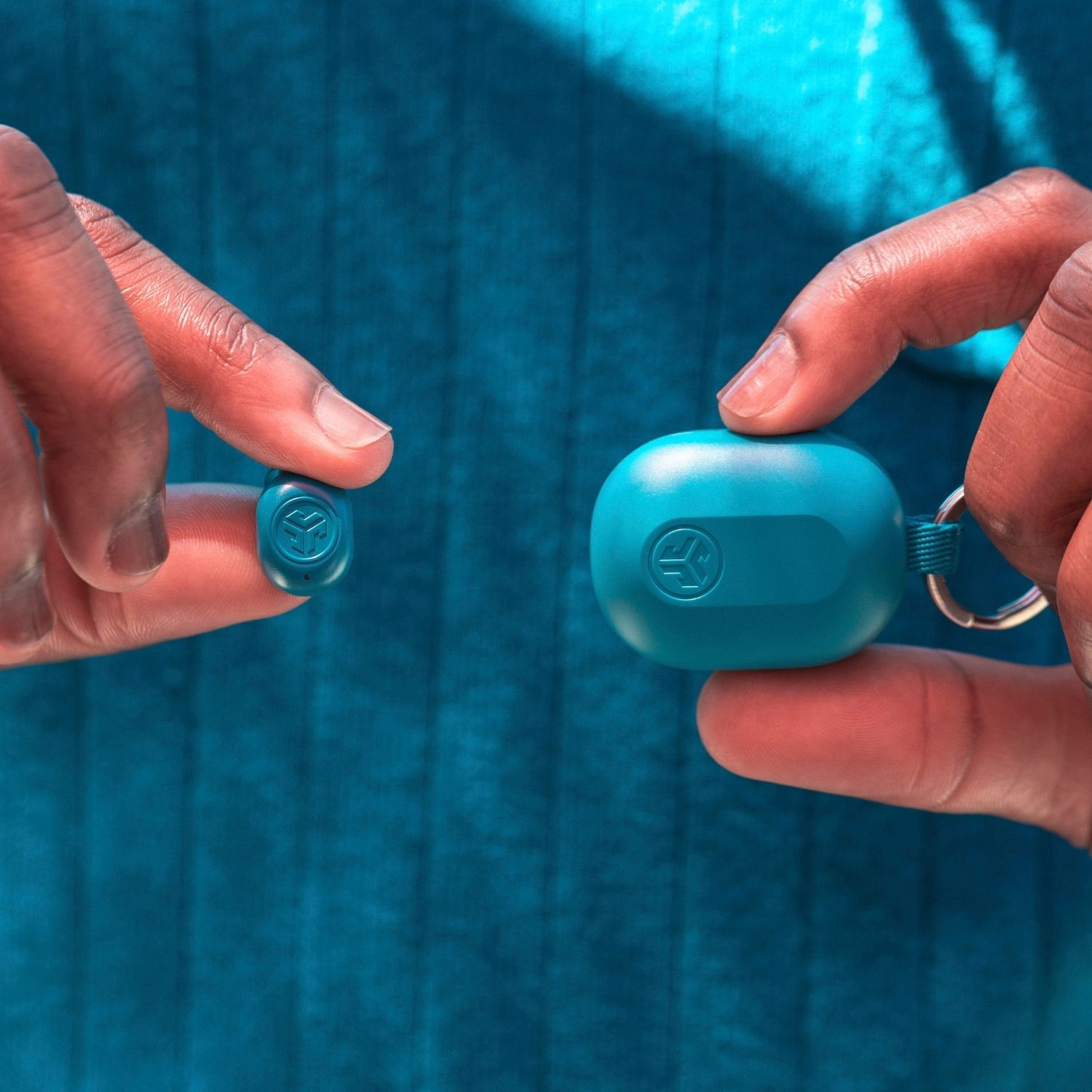 Manos sosteniendo auriculares y estuche de carga. Ambos turquesa, con logo. Fondo azul.