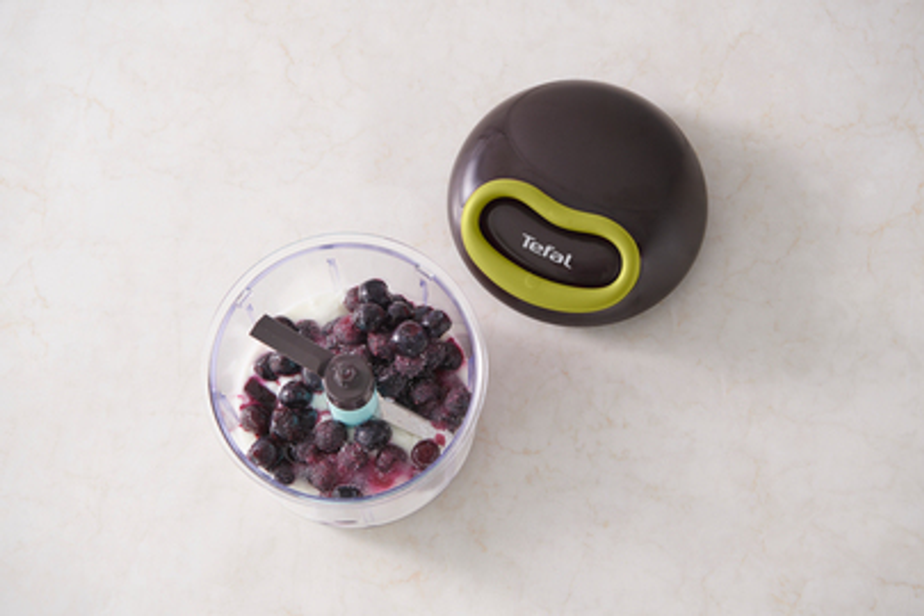 Ein Foodprozessor mit Blaubeeren und Joghurt, neben dem geschlossenen Deckel auf Marmor.