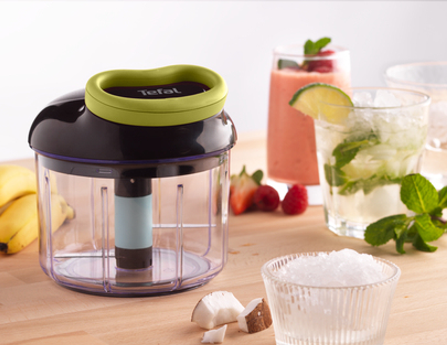 Ein Tefal-Zerkleinerer mit grünem Deckel, ein Glas mit Getränk und eine Schüssel mit Eis stehen auf Holz.