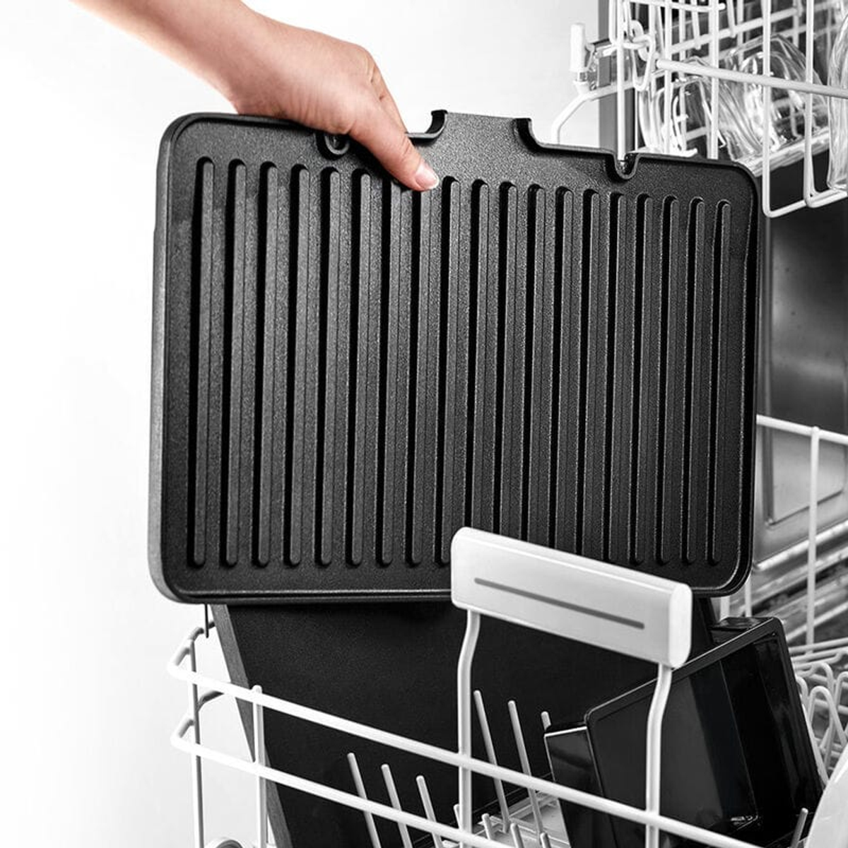 Ręka wyjmująca czarną patelnię grillową z kosza zmywarki z innymi przedmiotami.