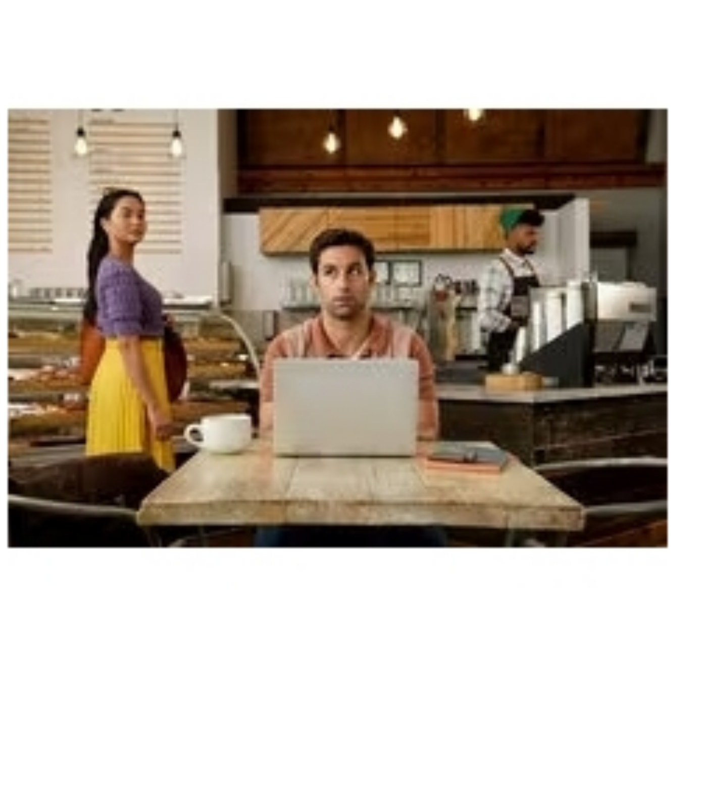 Ein Mann benutzt einen Laptop in einem Café, im Hintergrund eine Frau und ein Kellner.
