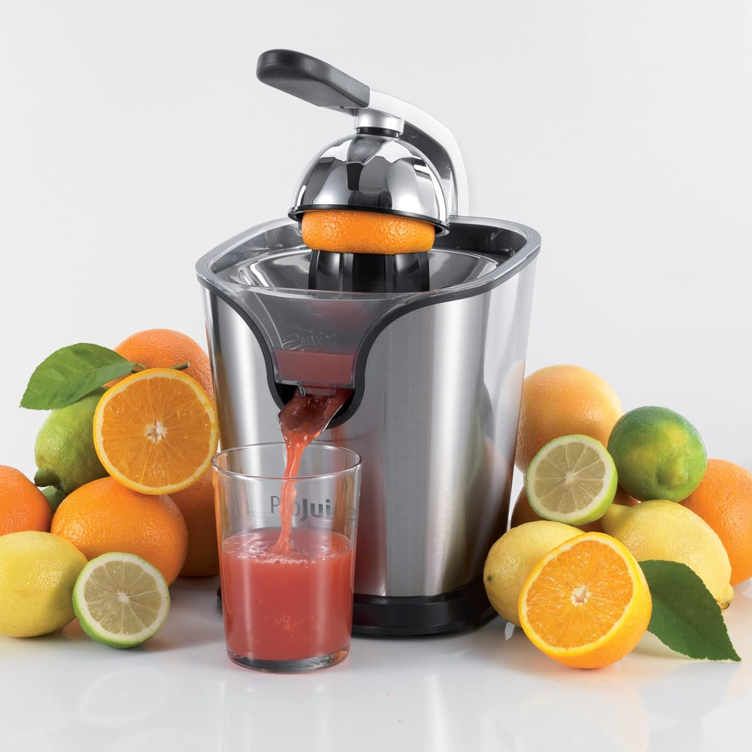 Una exprimidora está exprimiendo una naranja. Un vaso de jugo y frutas como limones y naranjas lo rodean.