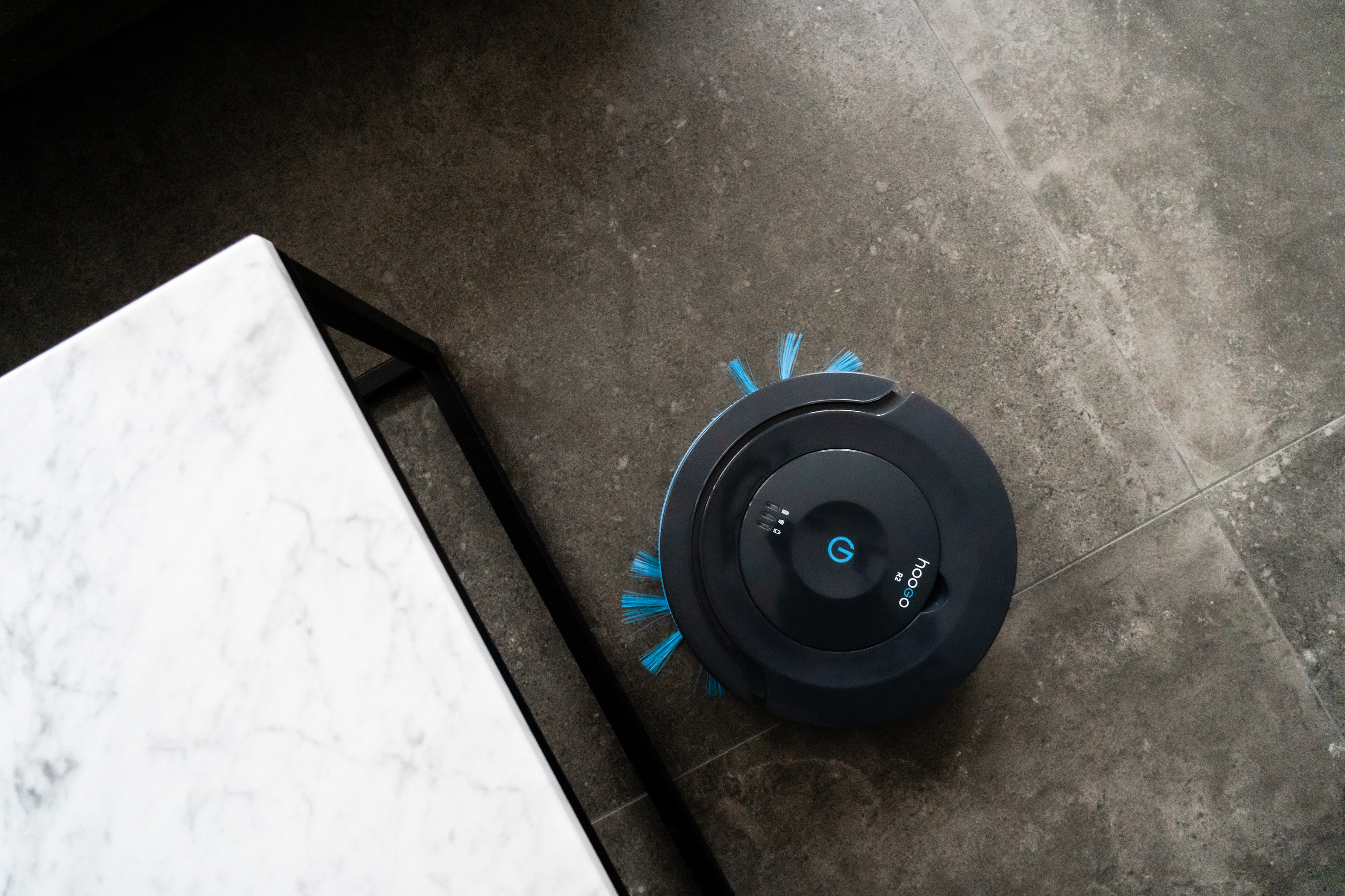 Ein schwarz-blauer Roboter-Staubsauger auf einem grauen Fliesenboden neben einem weißen Marmortisch.