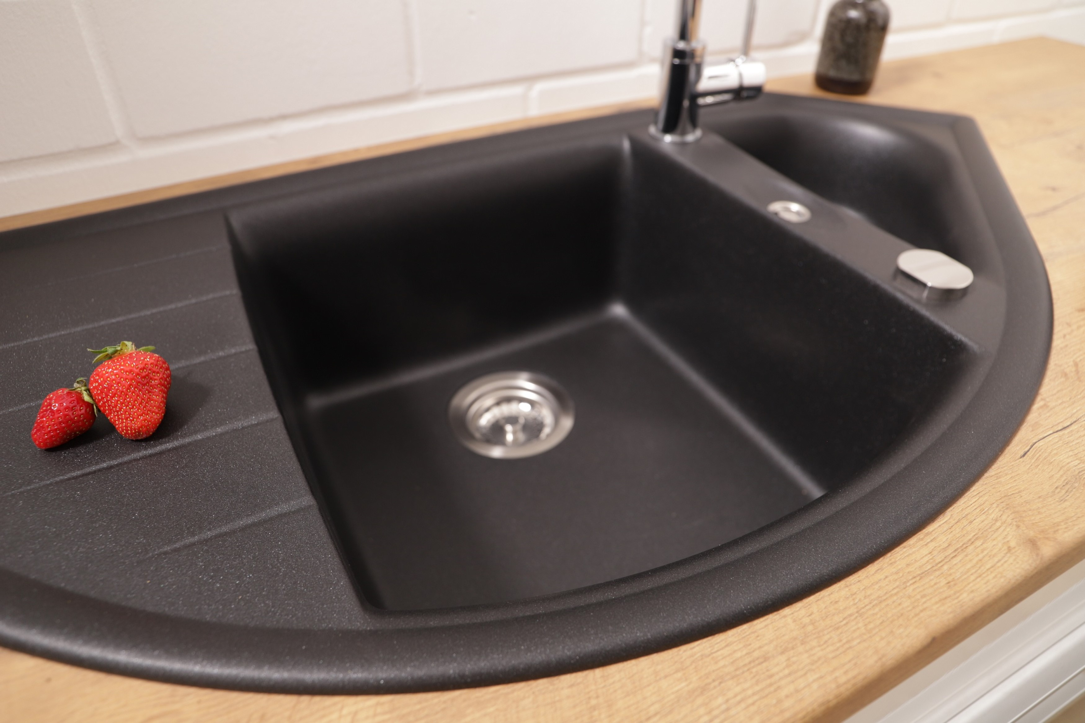 Ein schwarzes Küchenbecken mit einer Edelstahlarmatur. Zwei Erdbeeren liegen auf der Arbeitsplatte.