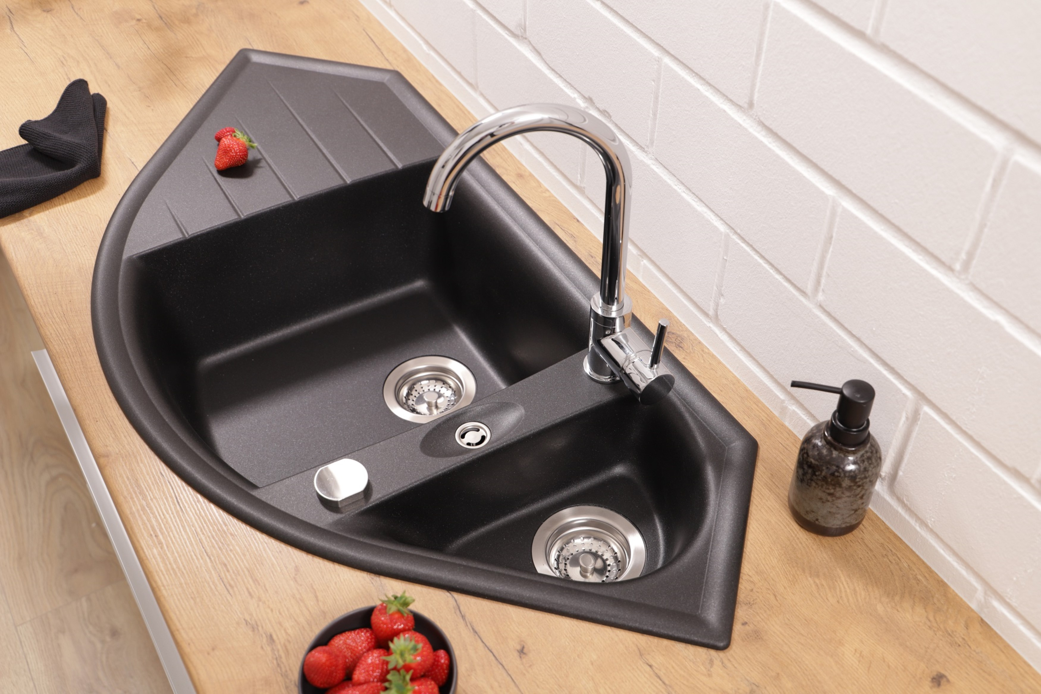 Schwarzes Eckwaschbecken mit Wasserhahn und Erdbeeren auf einer Holzplatte. Weiße Ziegelwand im Hintergrund.