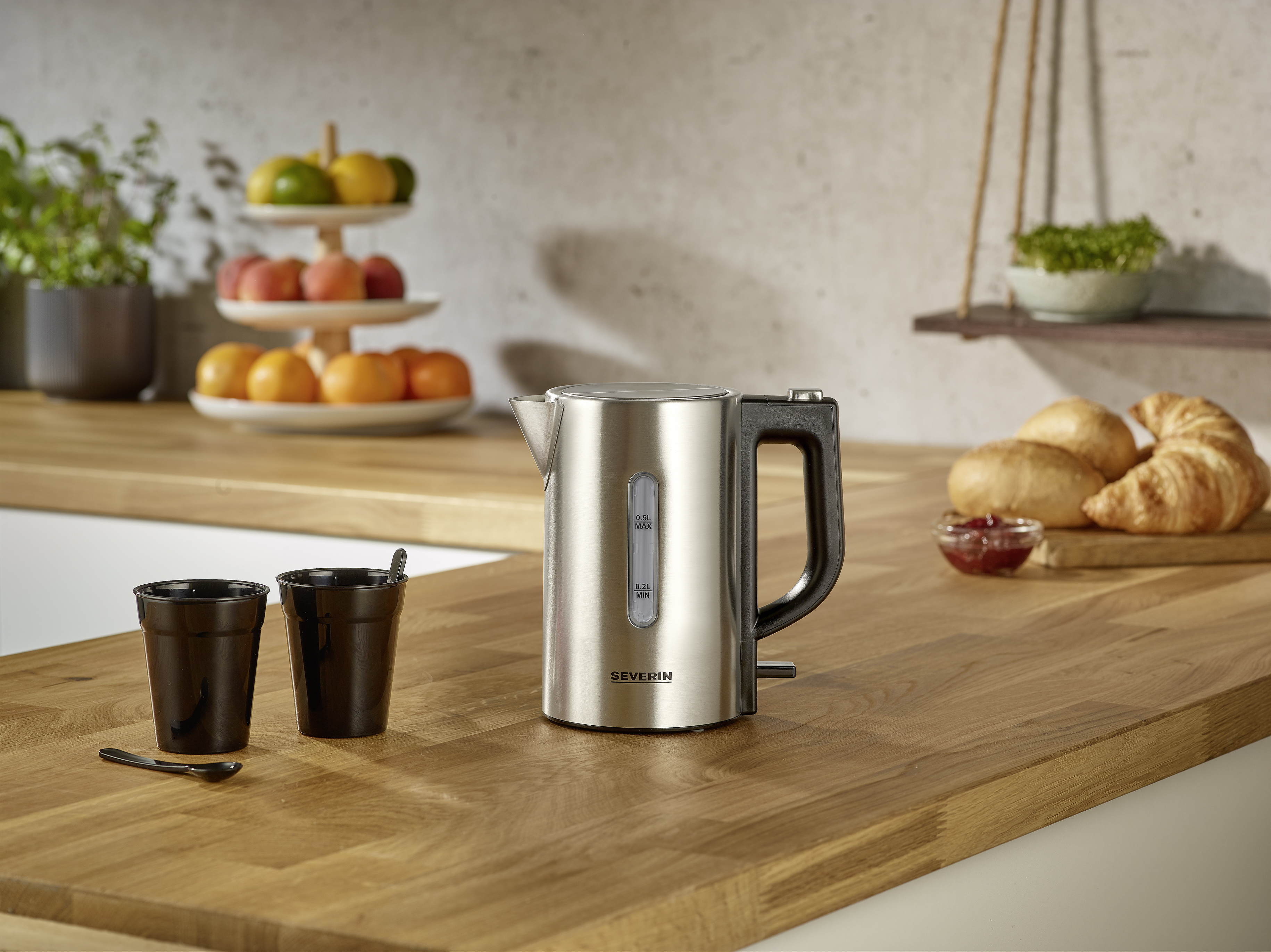 Czajnik elektryczny ze stali nierdzewnej na drewnianym blacie. W pobliżu są czarne filiżanki i łyżka.