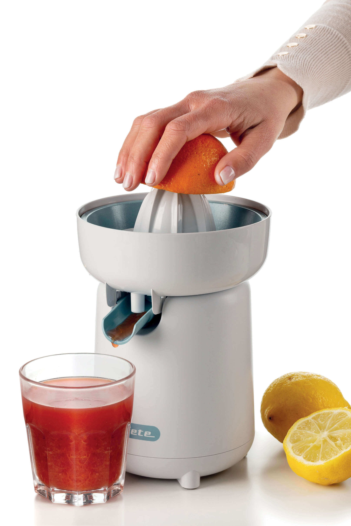 Eine Hand drückt eine Orange auf eine weiße Saftpresse. Ein Glas Saft und Zitronen sind ebenfalls vorhanden.