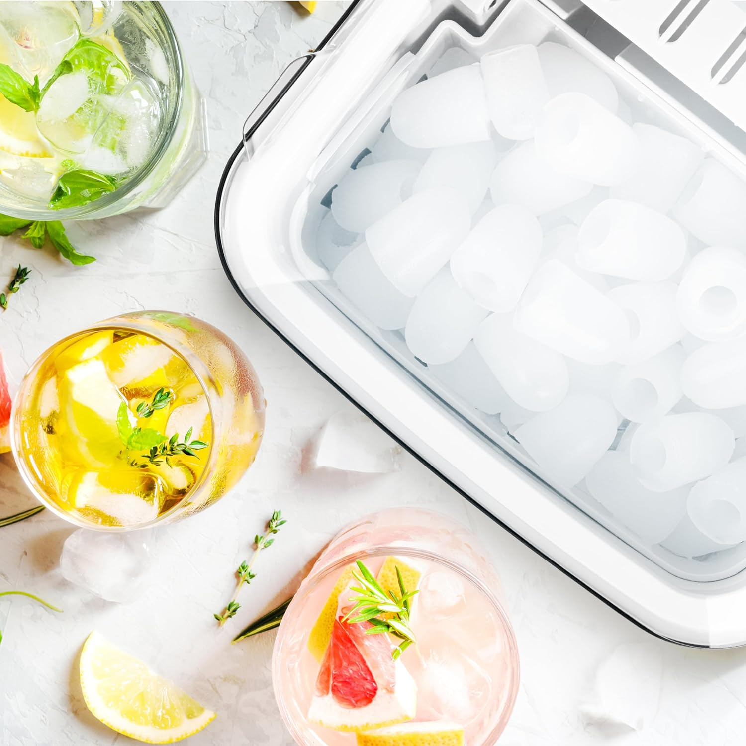 Cocktails mit Eiswürfeln, Zitronenscheiben und Kräutern auf weißer Oberfläche. Eismaschine im Hintergrund.