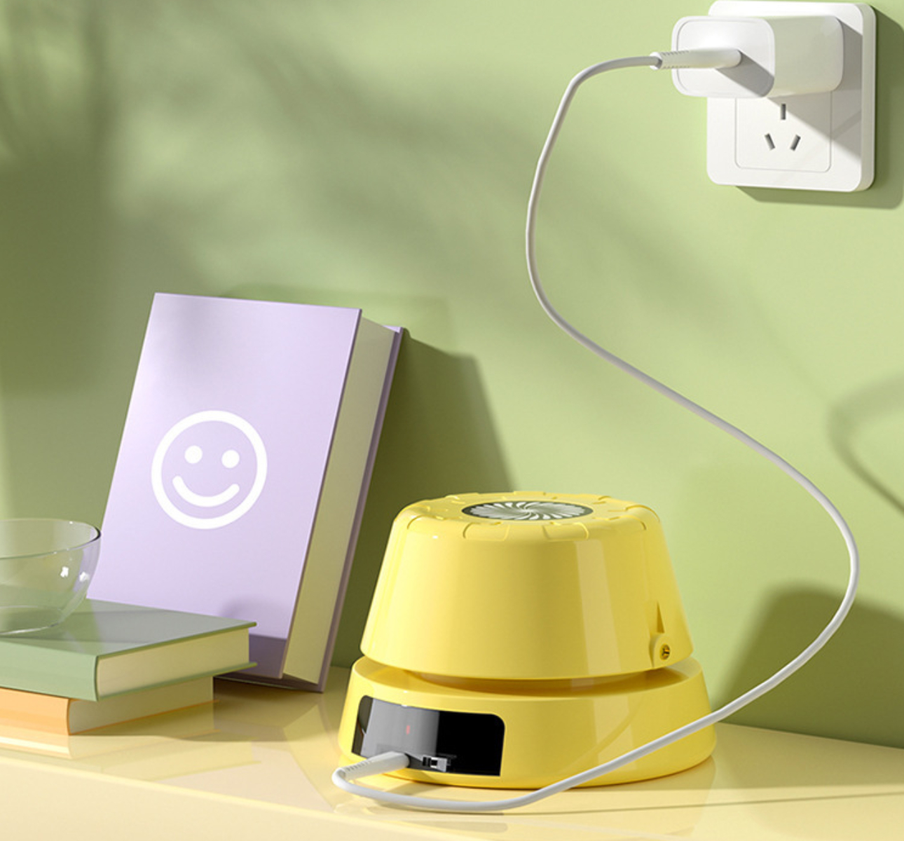 Gelber Ventilator mit Buch und Ladegerät. Der Ventilator ist eingesteckt. Weißer Hintergrund.