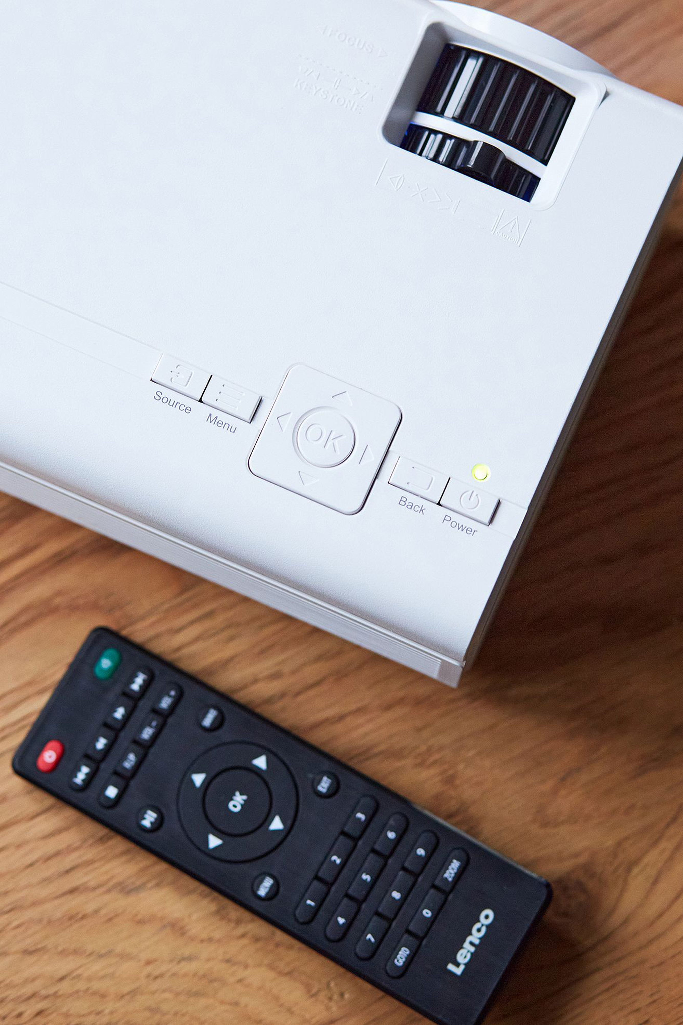 Biały projektor z pilotem na drewnianej powierzchni. Widoczne przyciski.