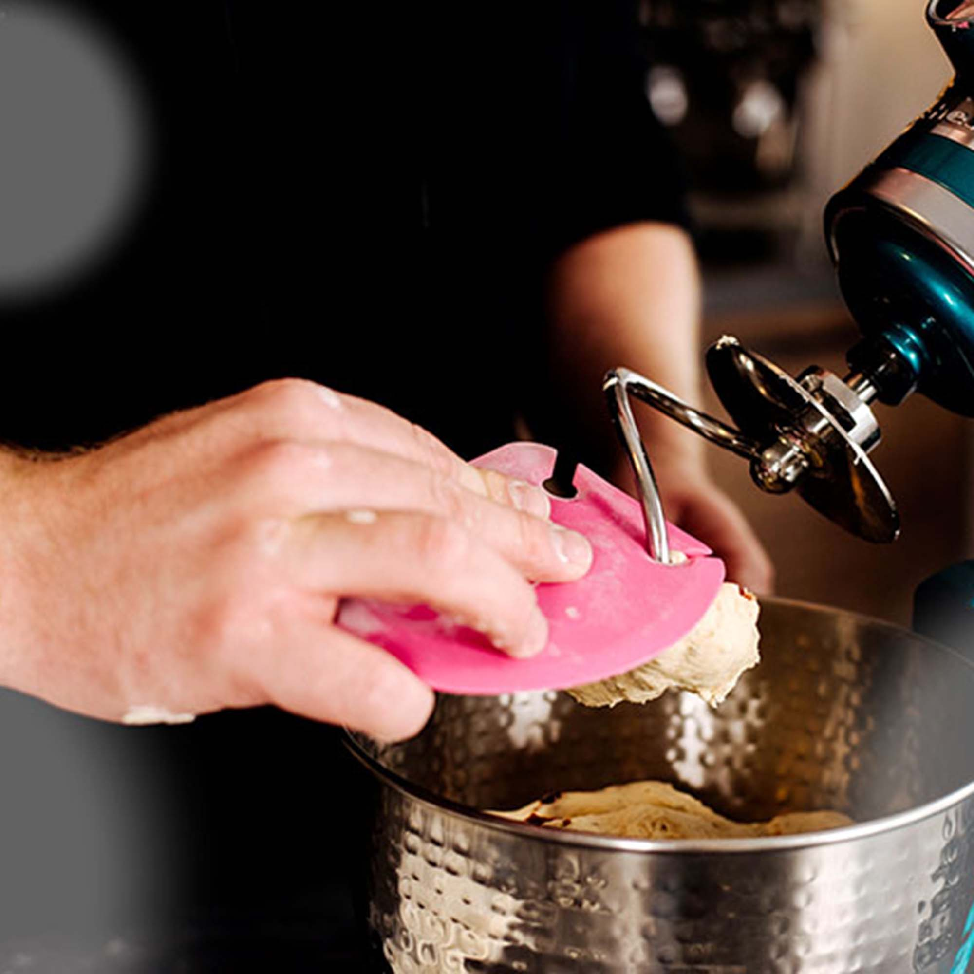 Eine Person verwendet einen pinkfarbenen Schaber, um Teig aus einer Mixerschüssel zu sammeln. Der Mixer ist teal.