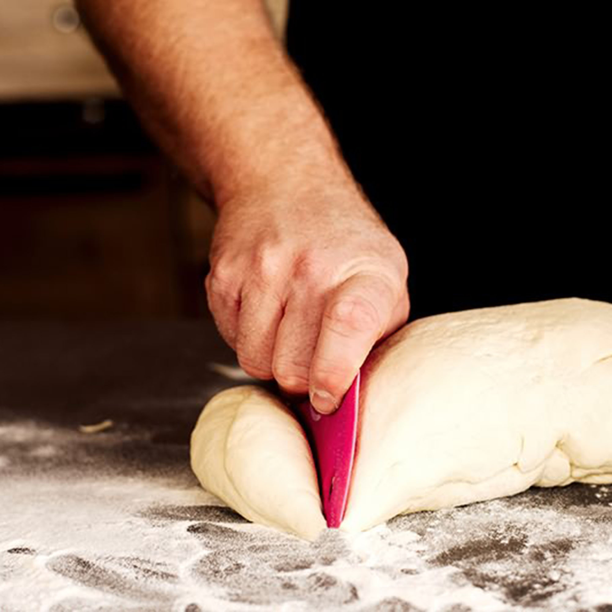 Eine Hand, die mit einem pinken Schaber Teig auf einer bemehlten Oberfläche schneidet.