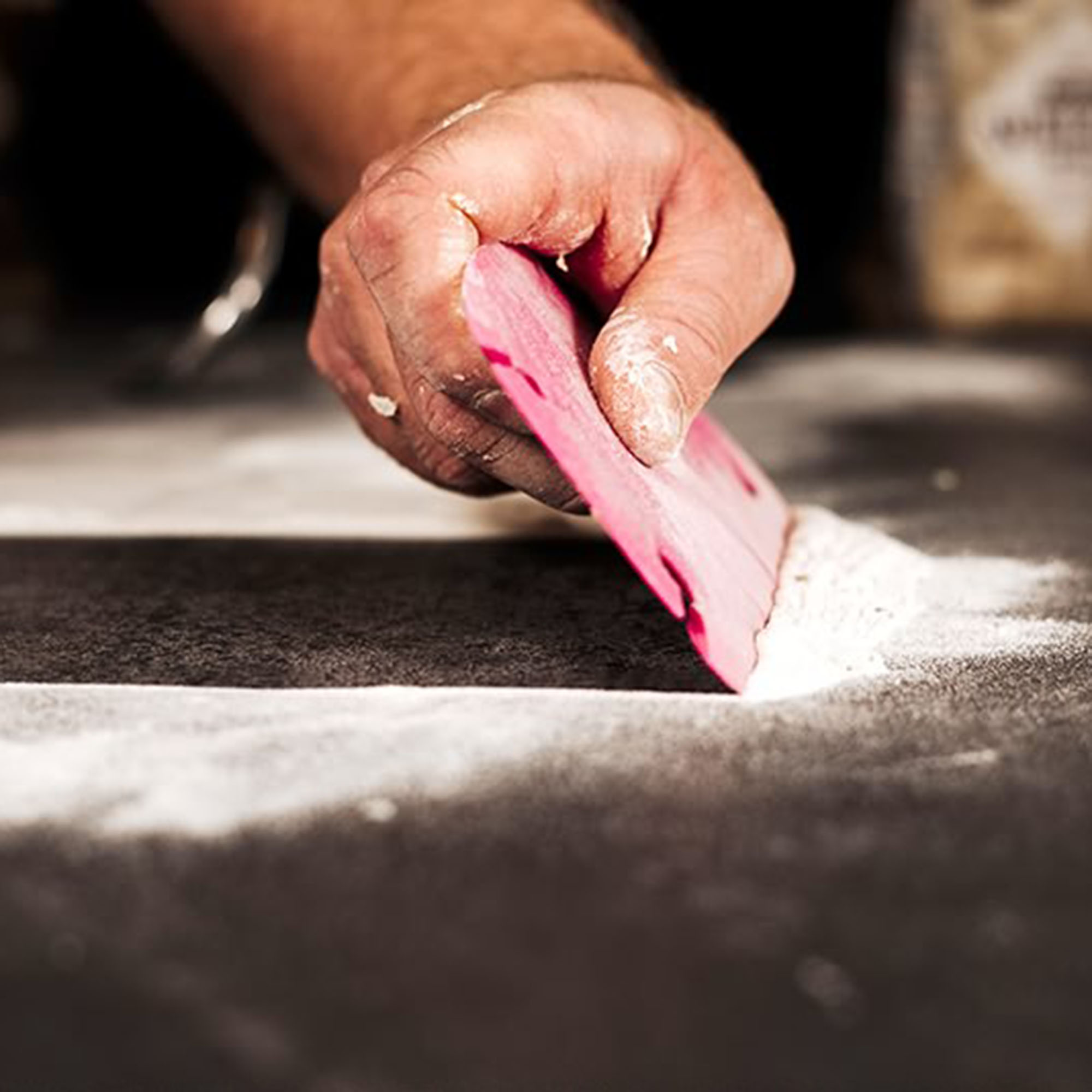 Eine Hand, die einen rosa Teigschaber auf einer schwarzen Oberfläche verwendet, um Mehl abzukratzen.