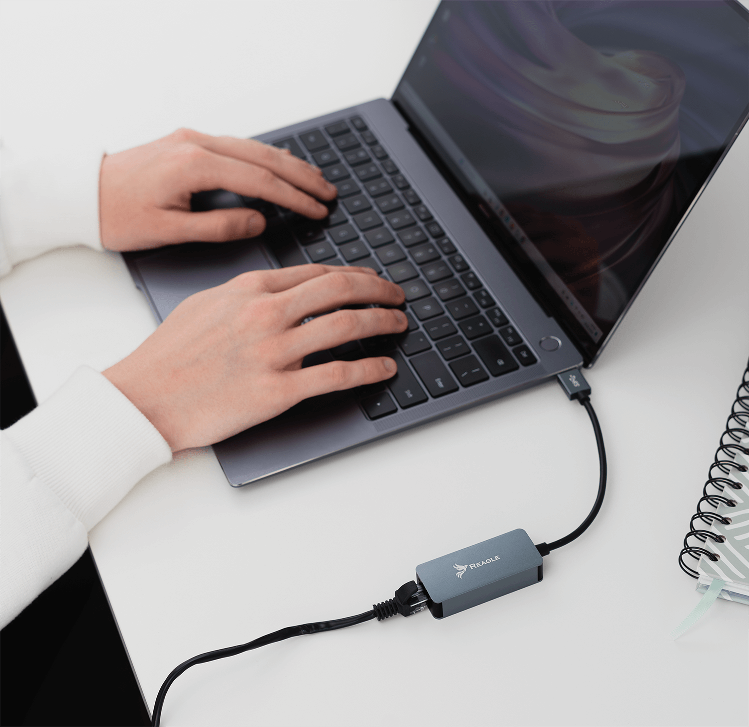 Osoba pisząca na szarym laptopie z podłączonym adapterem sieciowym. Białe biurko.