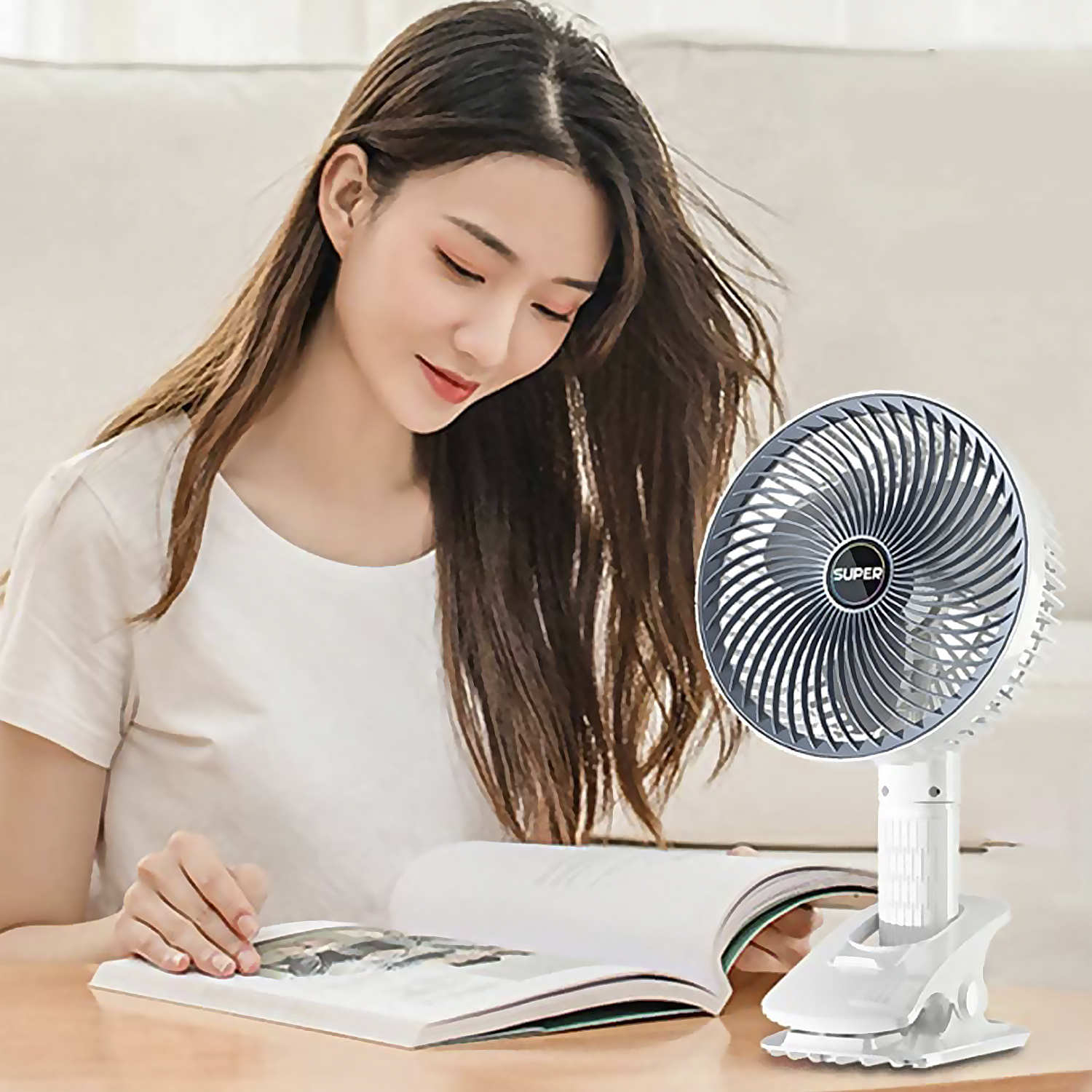 Una mujer leyendo un libro con un ventilador blanco sujetado a una mesa.