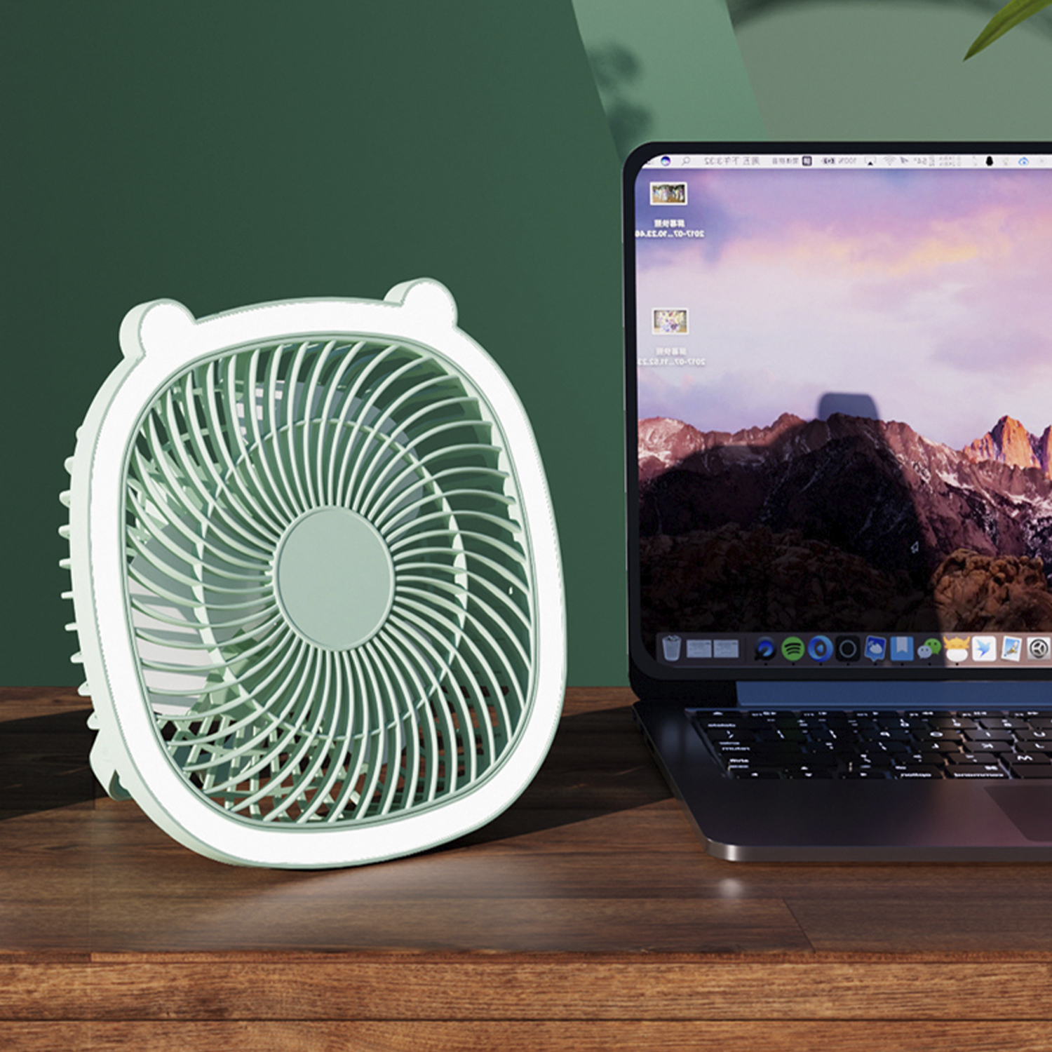 Piccolo ventilatore verde e laptop su un tavolo di legno. Il laptop mostra montagne.