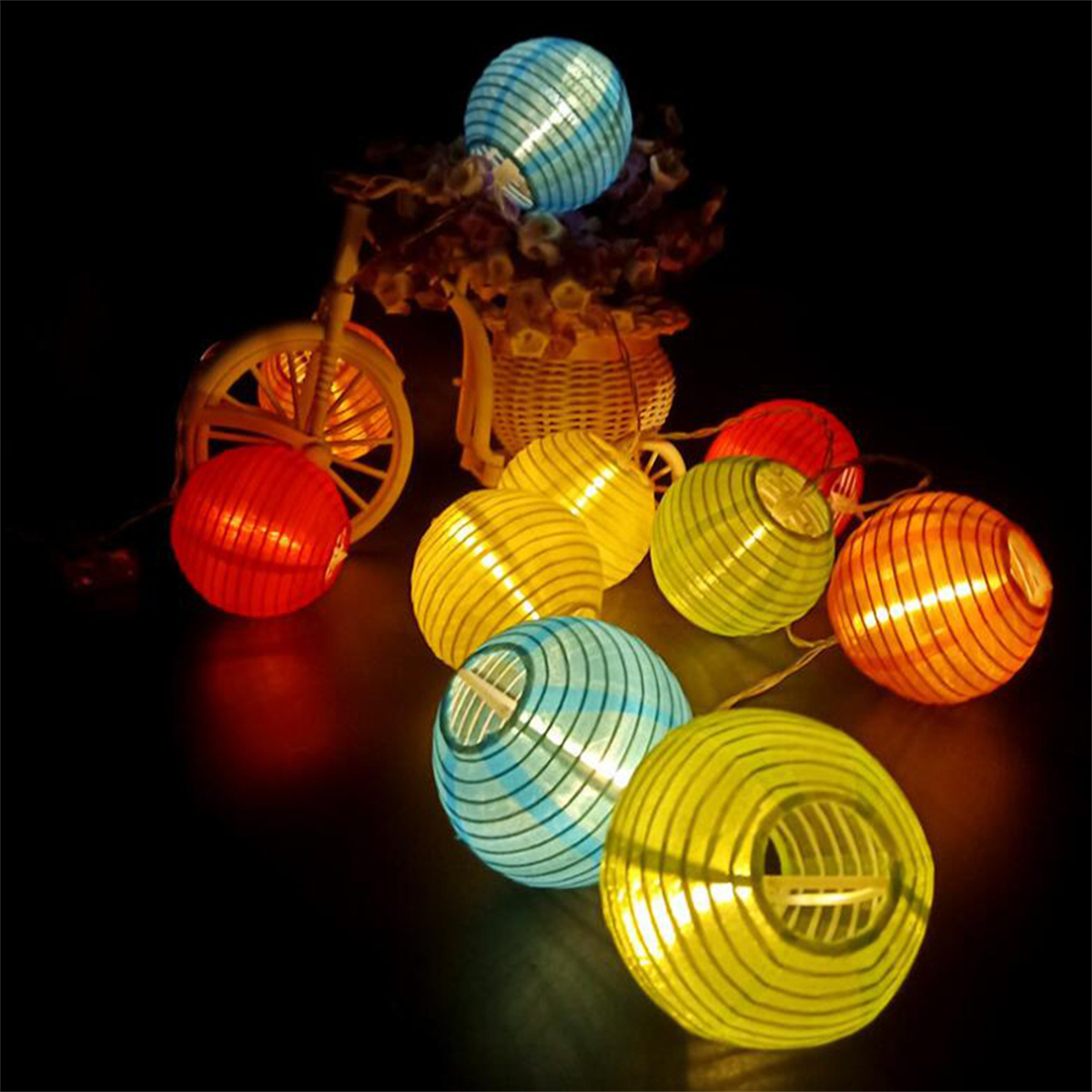 Lichterkette mit runden, bunten Laternen. Ein Fahrrad und Blumen im Hintergrund. Dunkle Umgebung.