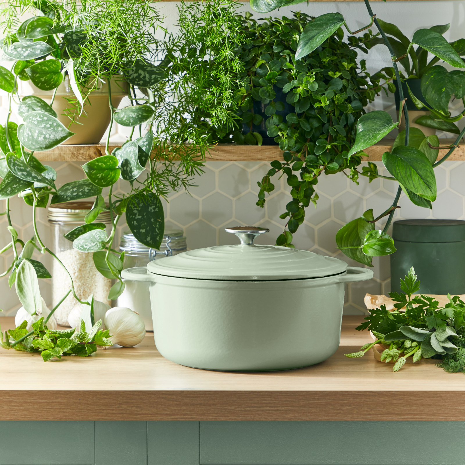 Zielony garnek z pokrywką stoi na drewnianym stole z roślinami i ziołami wokół niego. Tło stanowią rośliny.