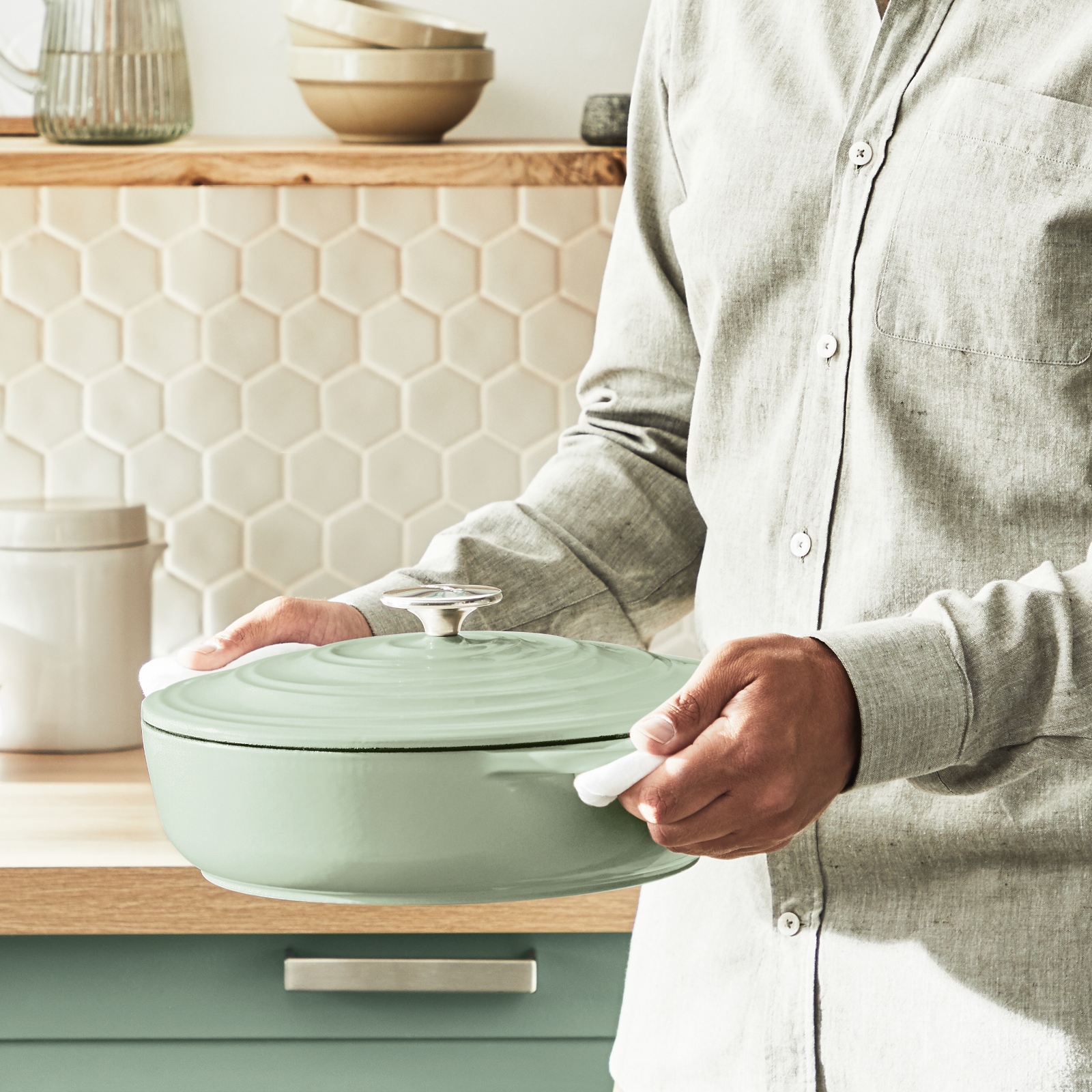 Osoba trzymająca jasnozielony żeliwny garnek z pokrywką. Otoczenie kuchni z drewnianymi blatami i białym tłem z płytek.