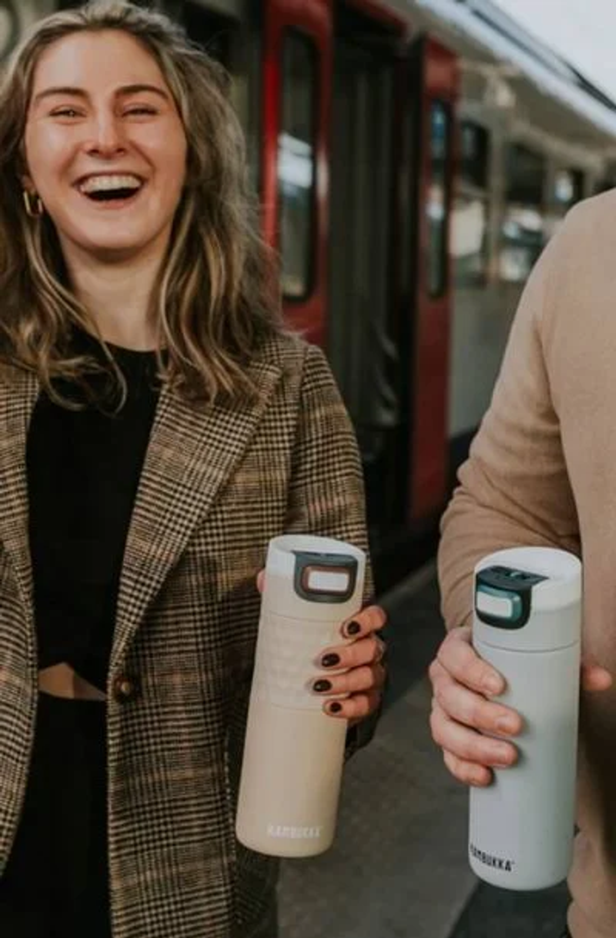 Zwei Personen halten Thermobecher. Eine lächelt, Bahnhofshintergrund. Beige und graue Becher.