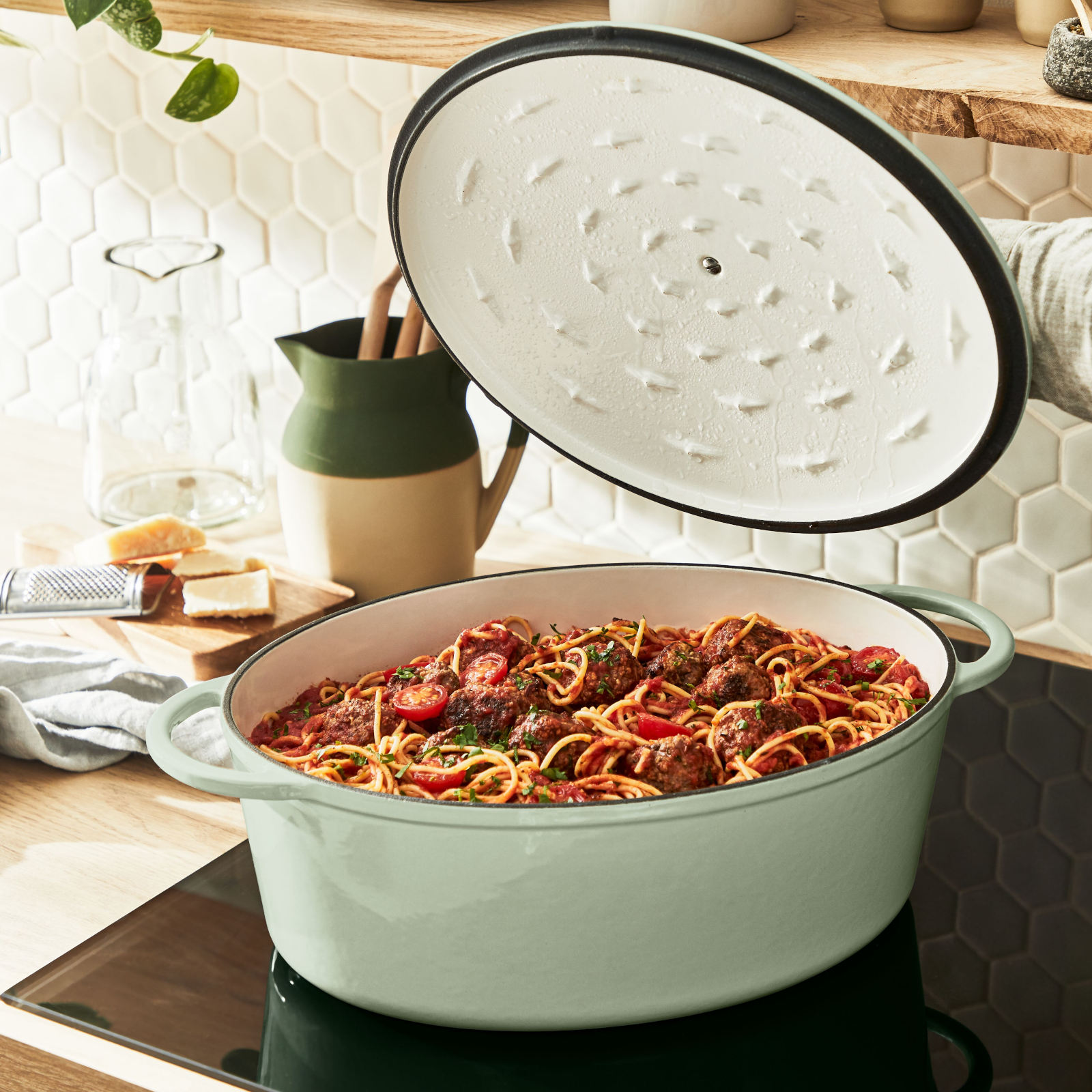Ein ovaler grüner Topf gefüllt mit Spaghetti und Fleischbällchen, mit offenem Deckel. Auf einem Herd.