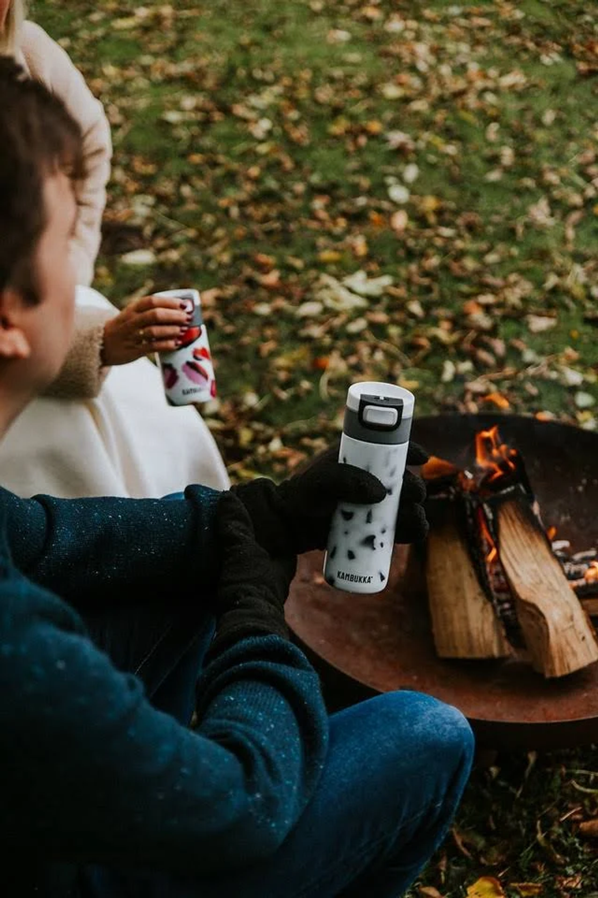 Zwei Personen sitzen im Freien mit Kaffeetassen in der Nähe einer Feuerstelle.
