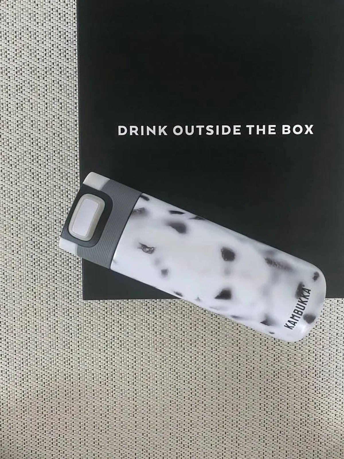 Ein weiß-schwarz gefleckter Reisebecher auf schwarzem Untergrund mit dem Text 'DRINK OUTSIDE THE BOX' darüber.
