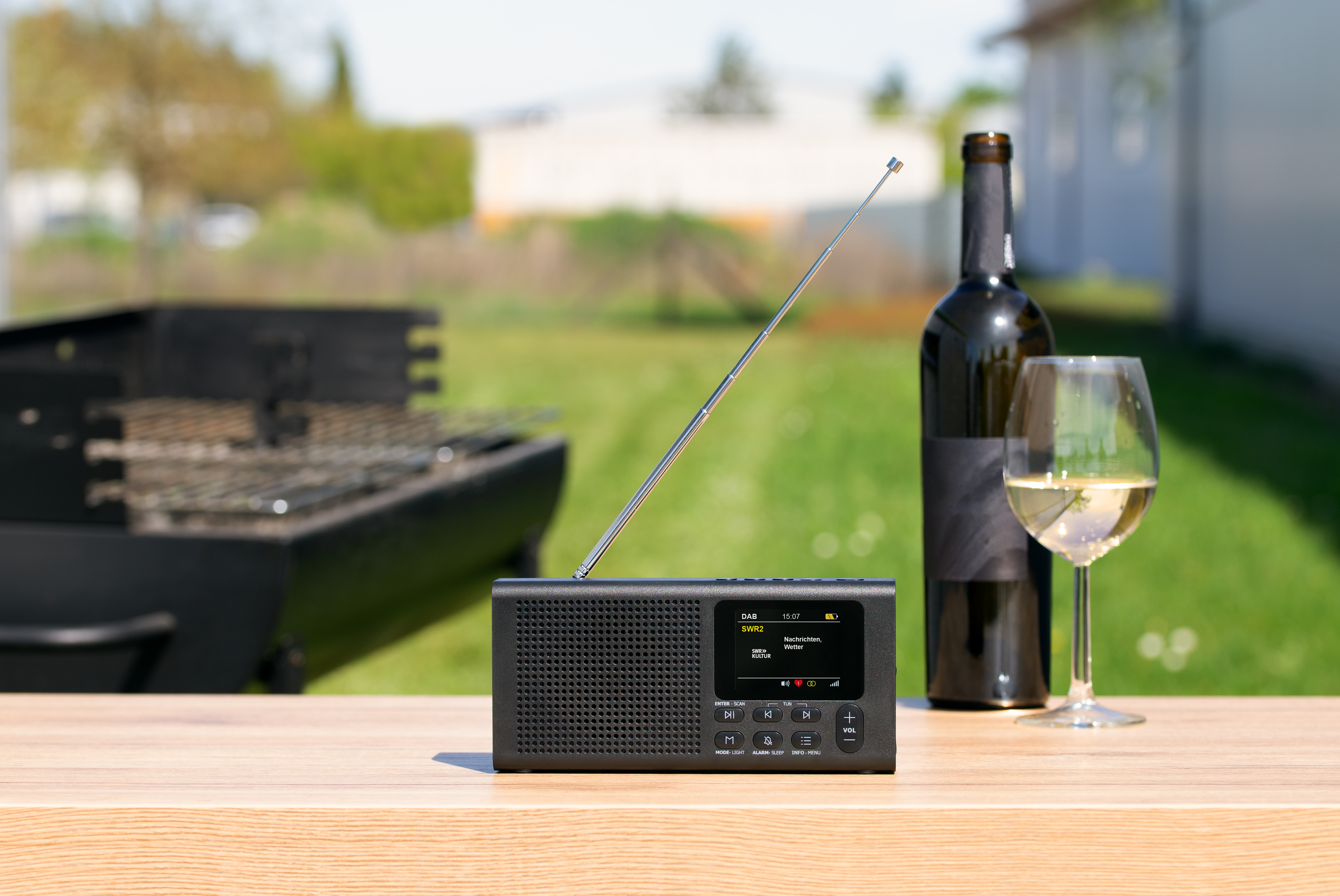 Ein schwarzes Radio mit Antenne, eine Flasche Wein und ein Weinglas auf einem Holztisch im Freien.