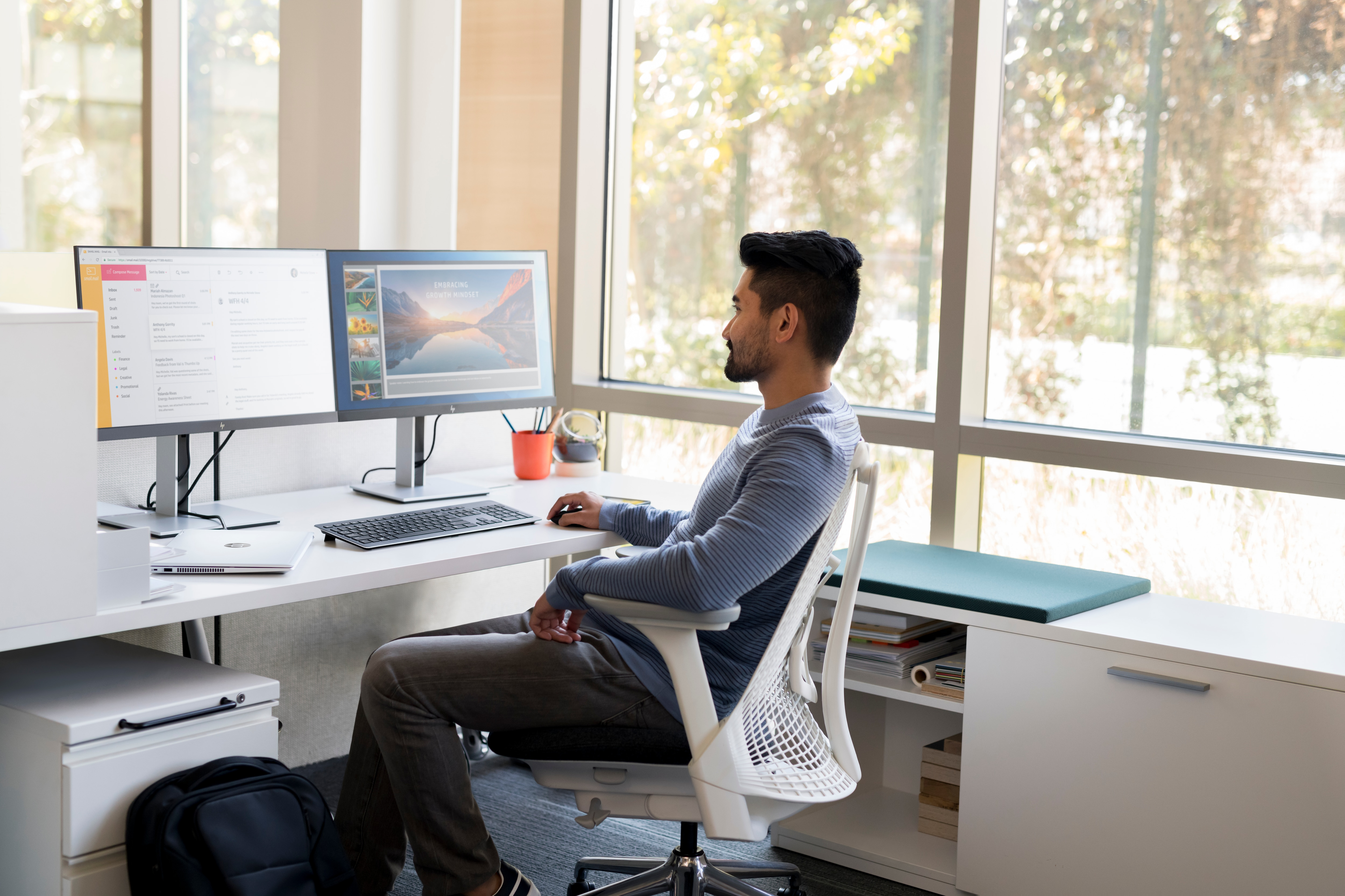 Man sitting at a desk with two monitors. Background is window with outdoor view.